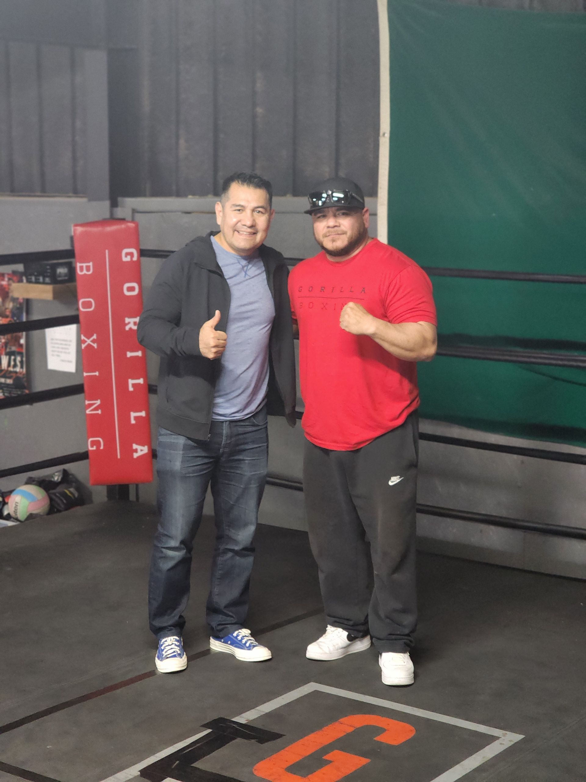 Two men smiling and giving thumbs up in a boxing gym, standing by a ring and red corner padding.
