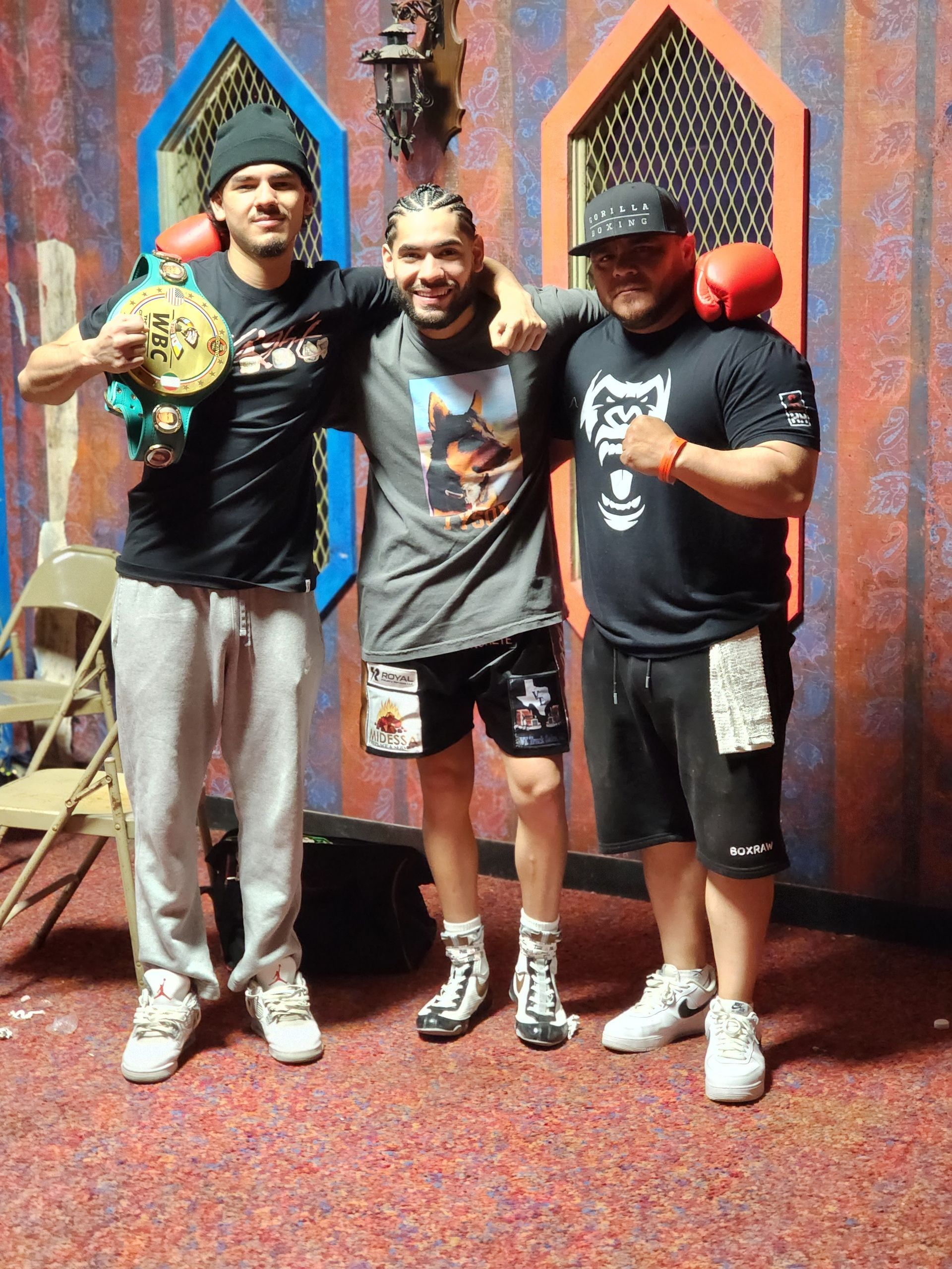 Three men posing together indoors, smiling, with championship belts and awards in front of a colorful door.