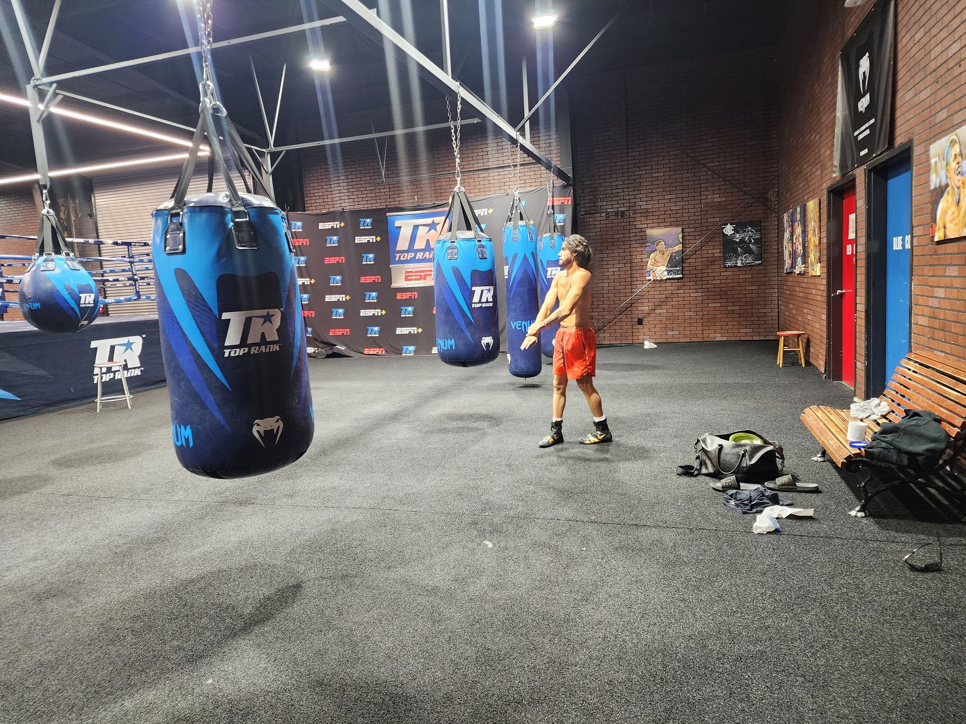 Person in orange shorts standing in a boxing gym beside hanging heavy bags.