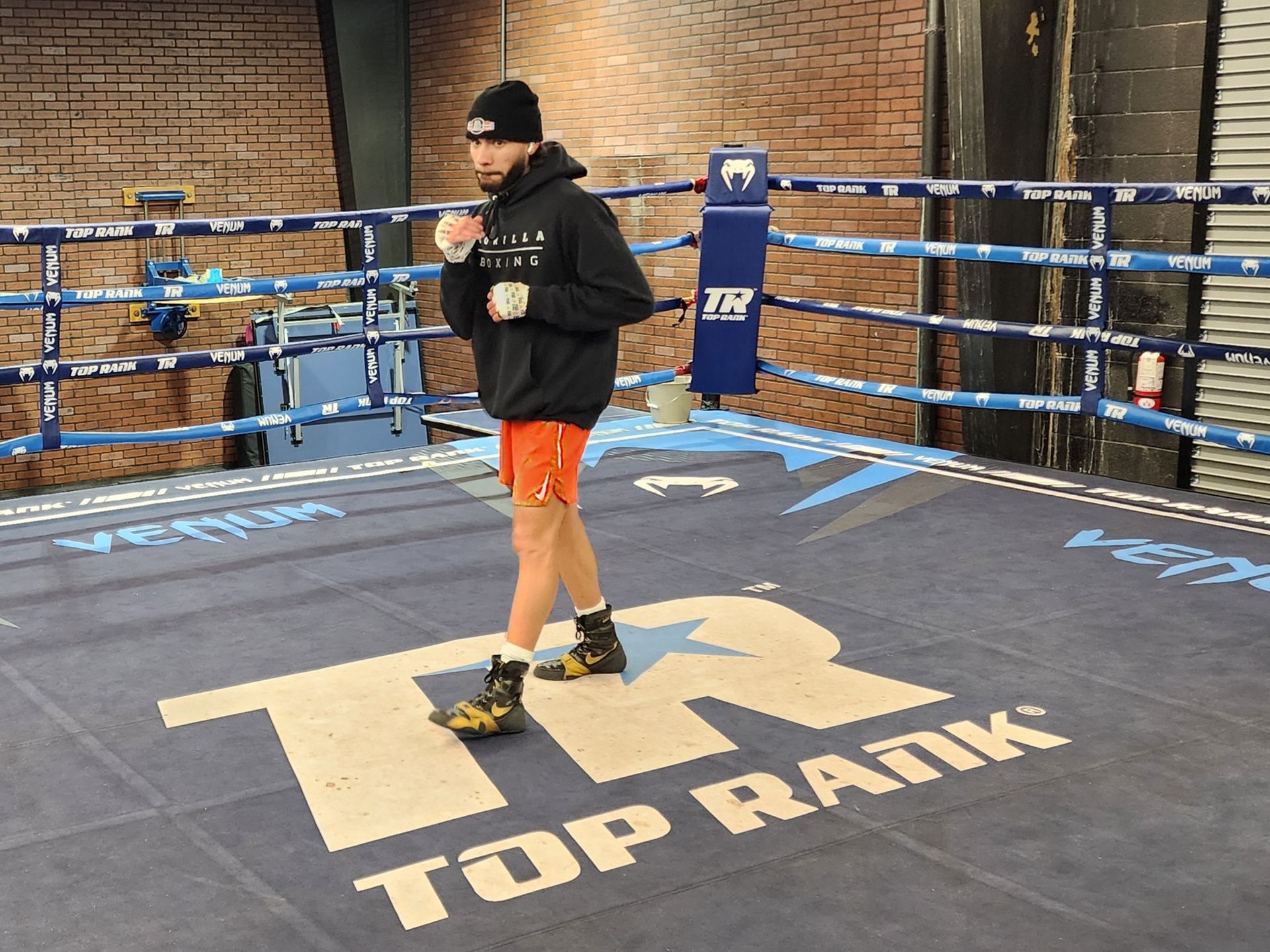 Person standing on a boxing ring mat with Top Rank logo, wearing black hoodie and orange shorts.