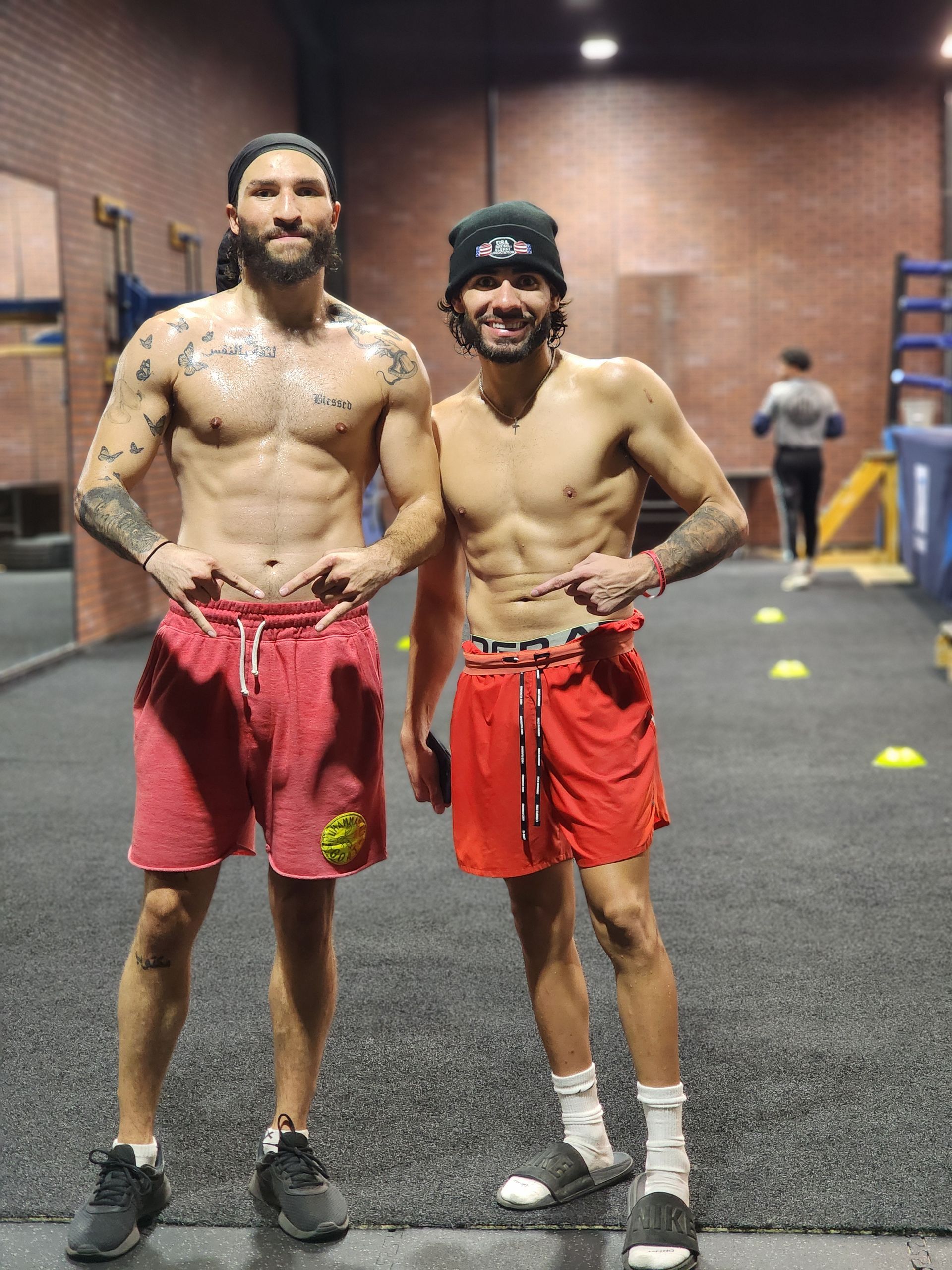 Two shirtless athletes in red shorts posing in a gym, one with tattoos and both smiling.