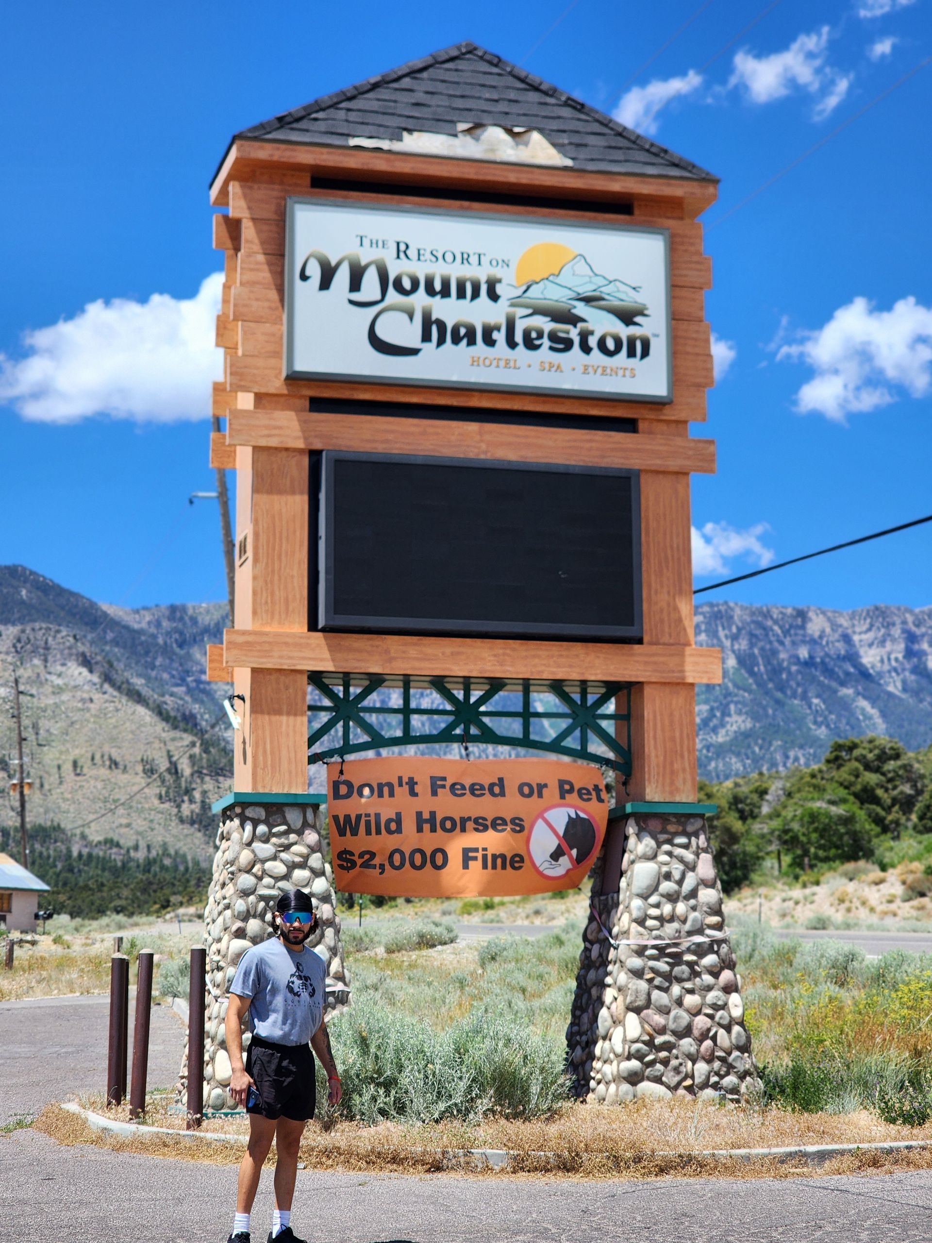 Sign for Mount Charleston with a warning about wild horses and fines, beside a person on a roadside.