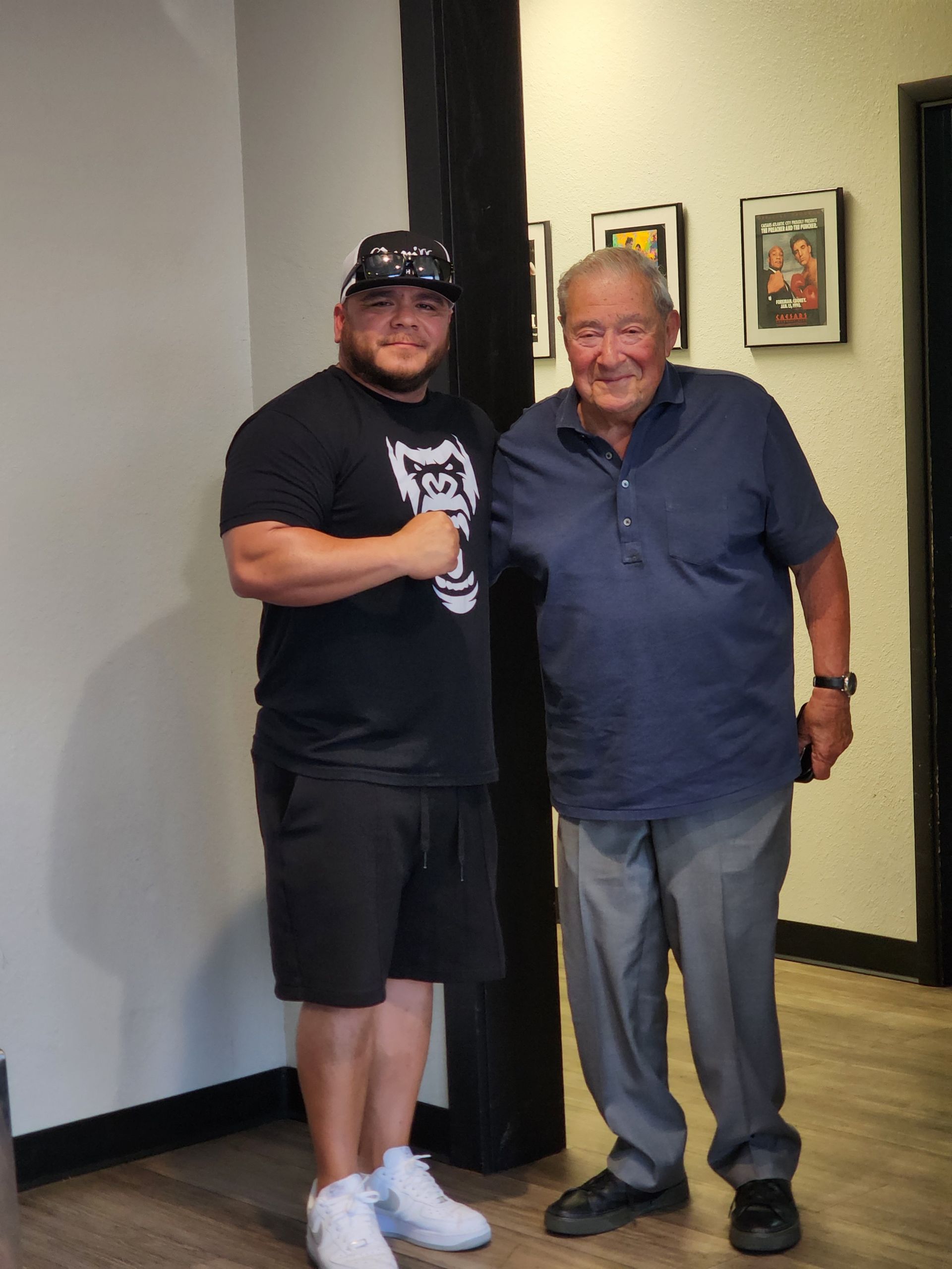 Two smiling men stand together indoors by a black column, one holding a drink.
