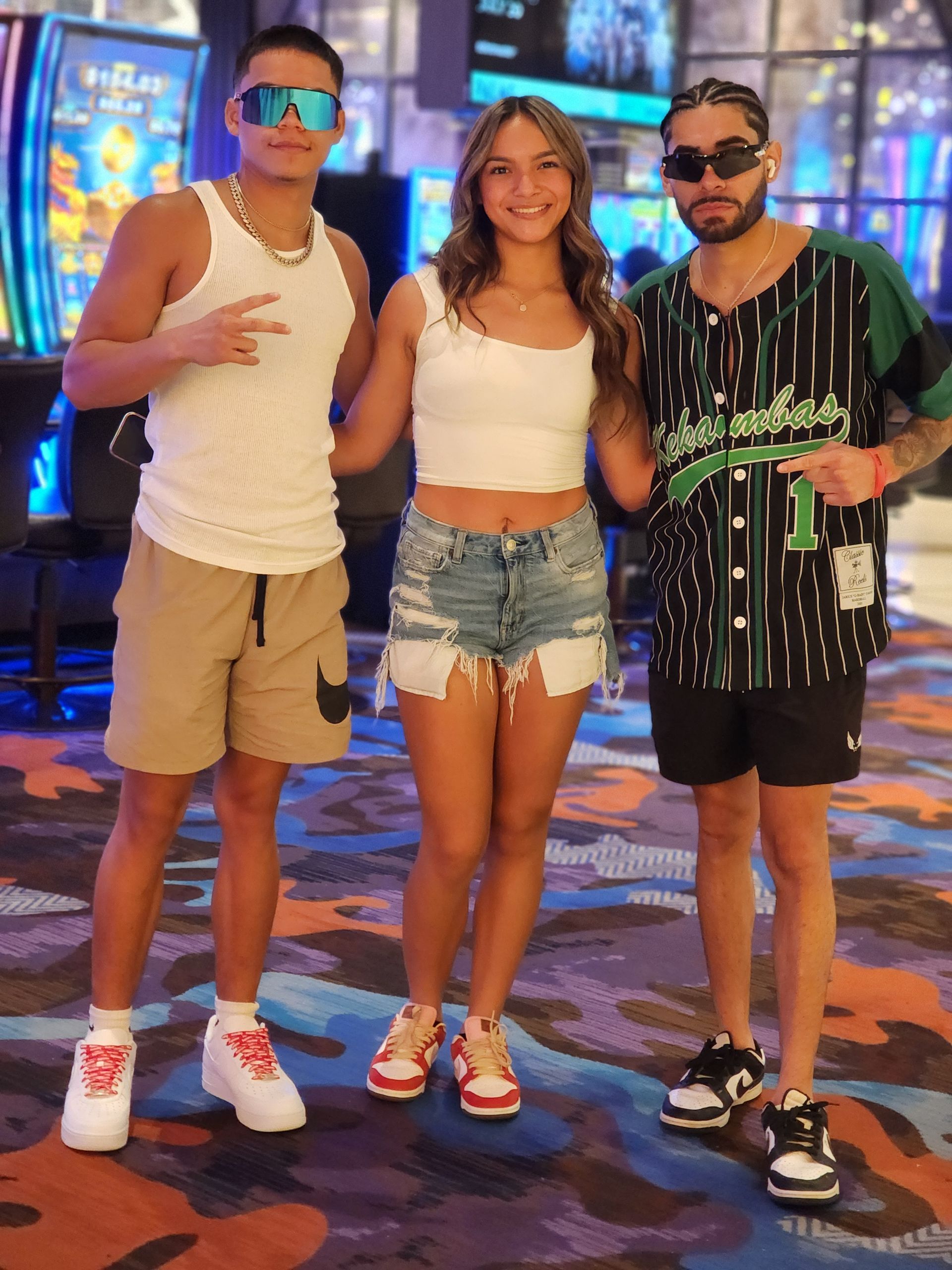 Three people posing in a neon-lit arcade, smiling and wearing casual summer clothes.