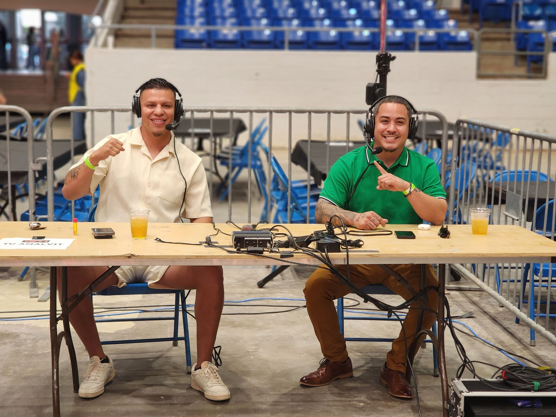 Two men wearing headsets sit at a table in an indoor arena, speaking into microphones.