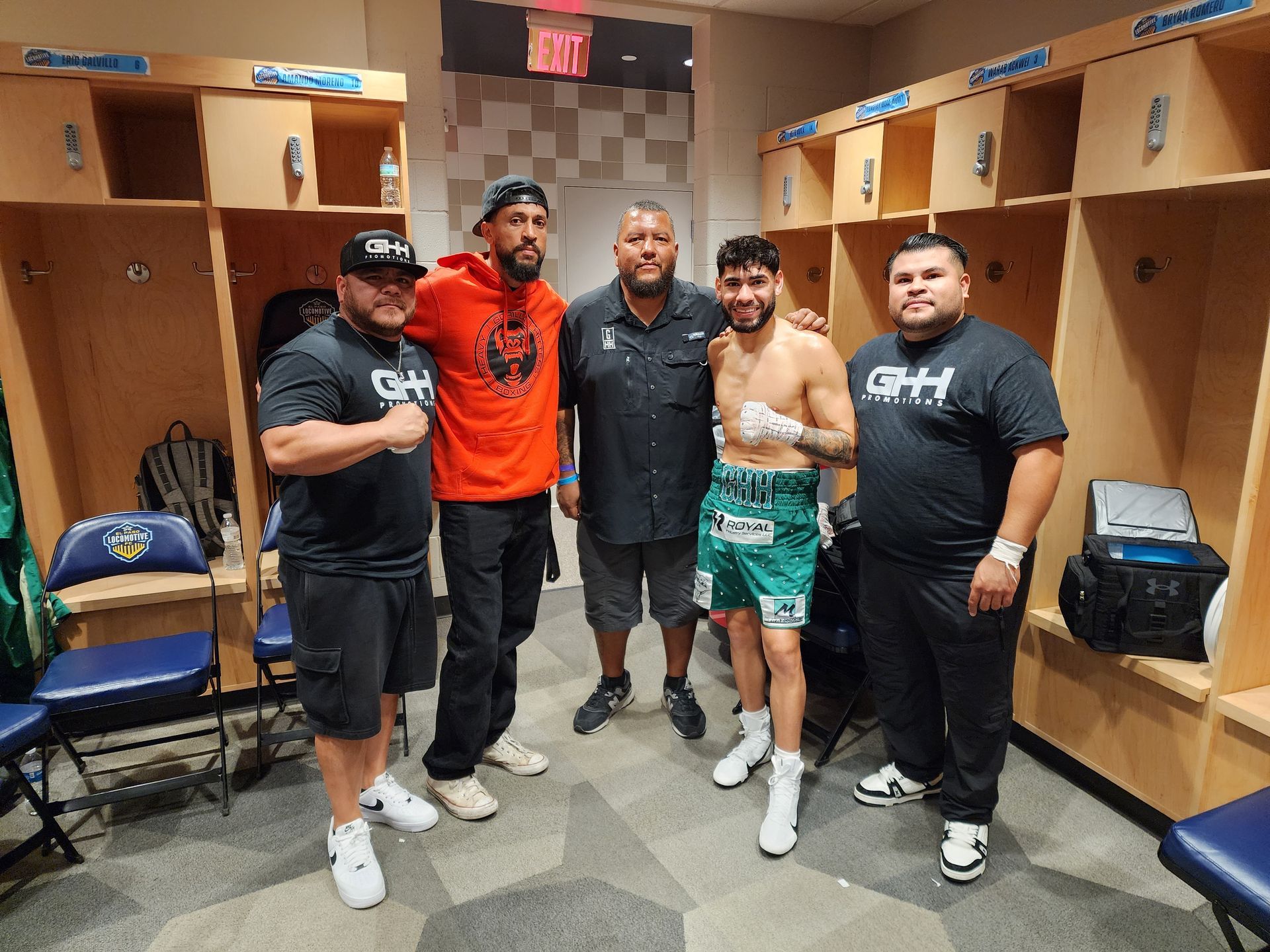 Five men pose in a locker room, one shirtless with a championship belt and others in team gear.
