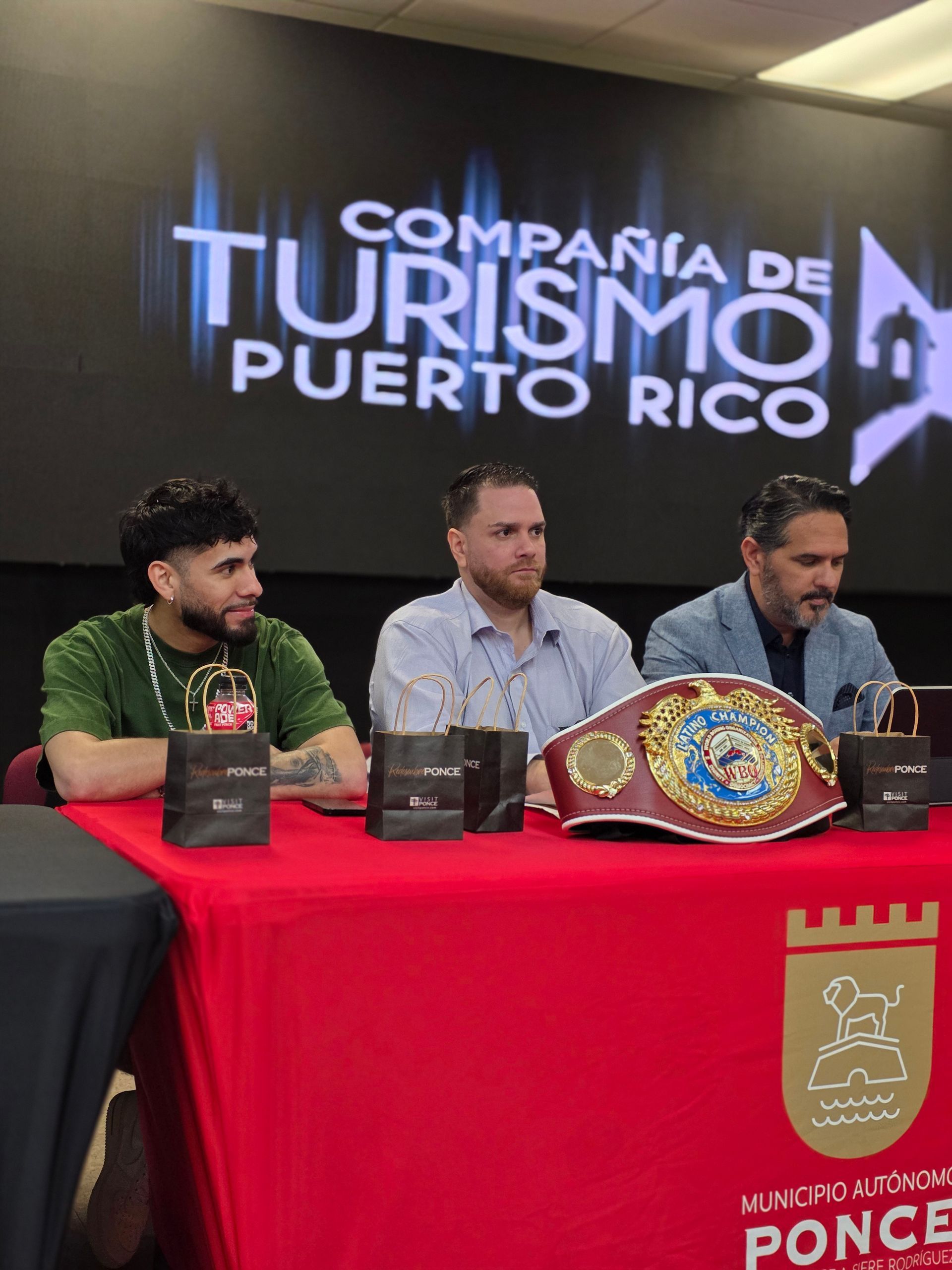 Three men at a red-covered press table with a tourism sign and championship belt behind them.