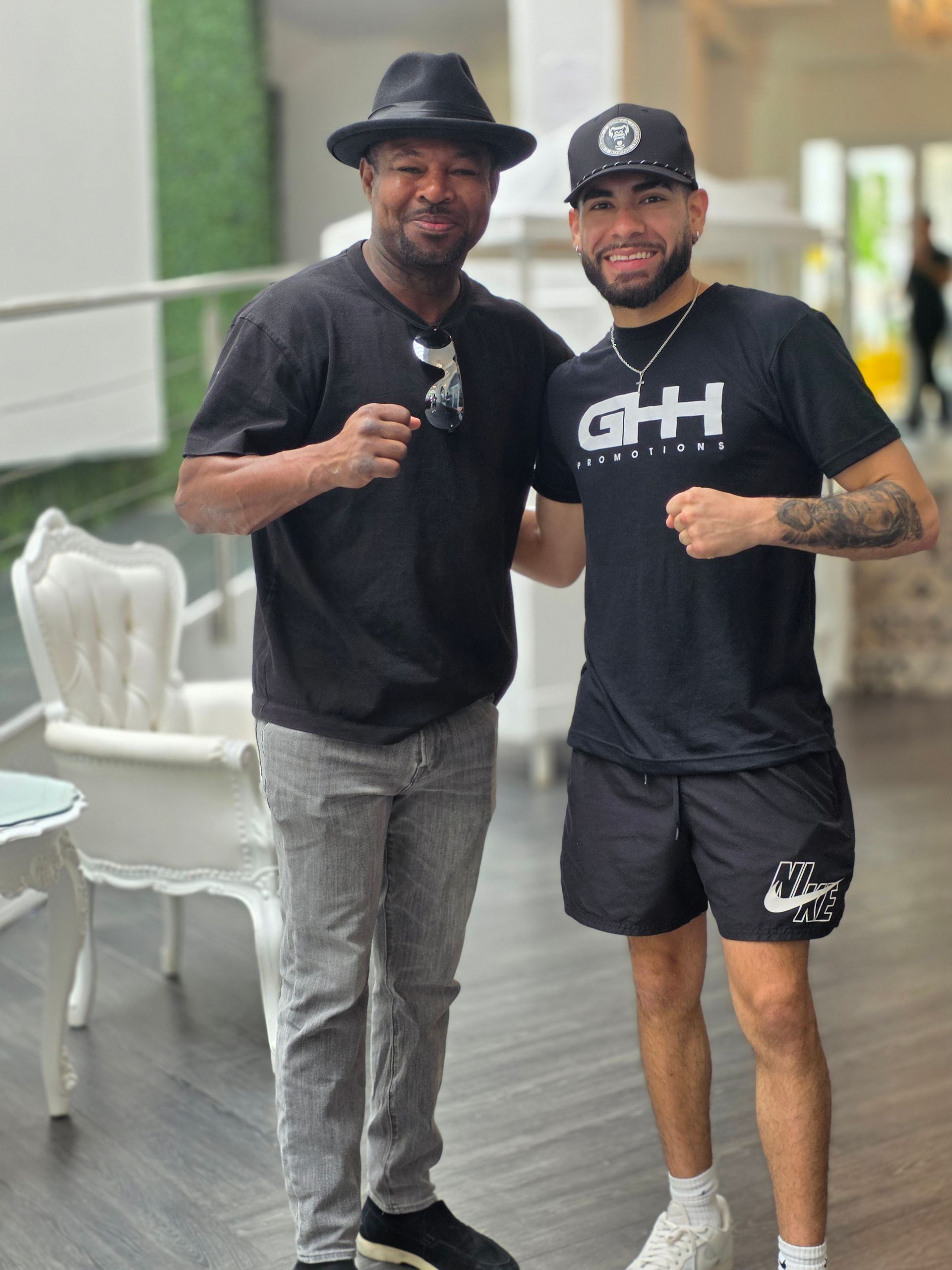 Two men pose smiling indoors, both in black shirts and shorts, one wearing a hat.