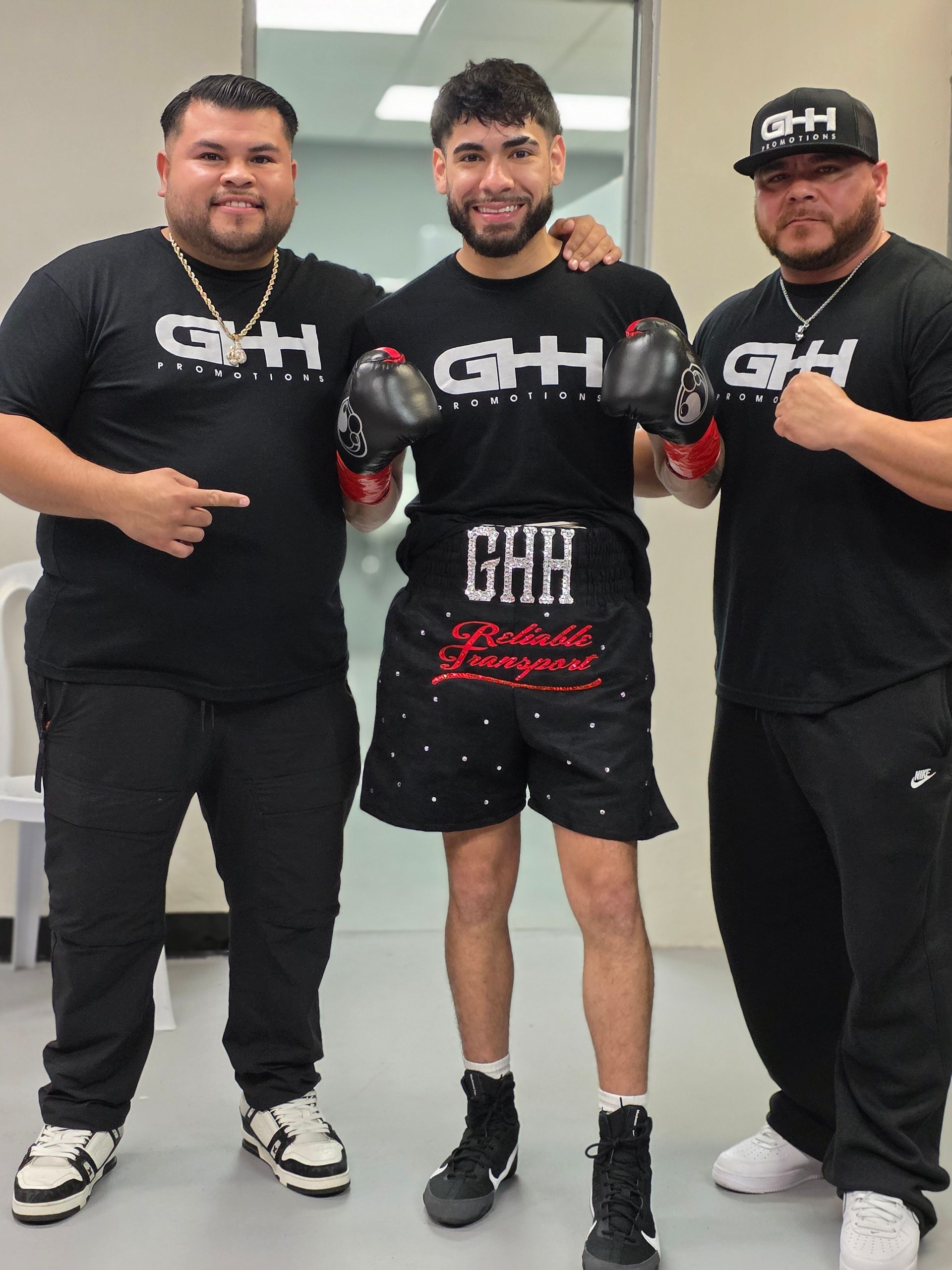 Three men in black GH shirts posing indoors; center wears boxing gloves and shorts.