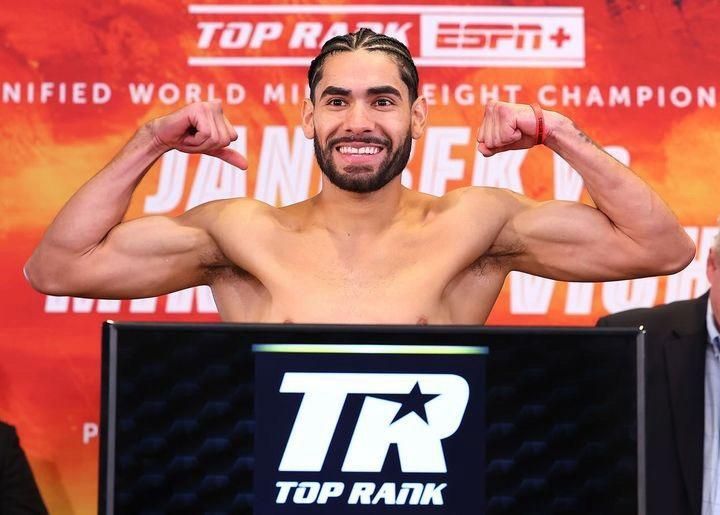 Smiling boxer flexing at a Top Rank weigh-in with a red ESPN+ championship backdrop