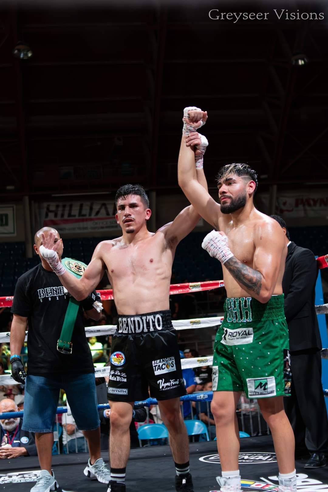Two boxers in the ring, one in black shorts and one in green, raise their arms as the referee stands behind them.