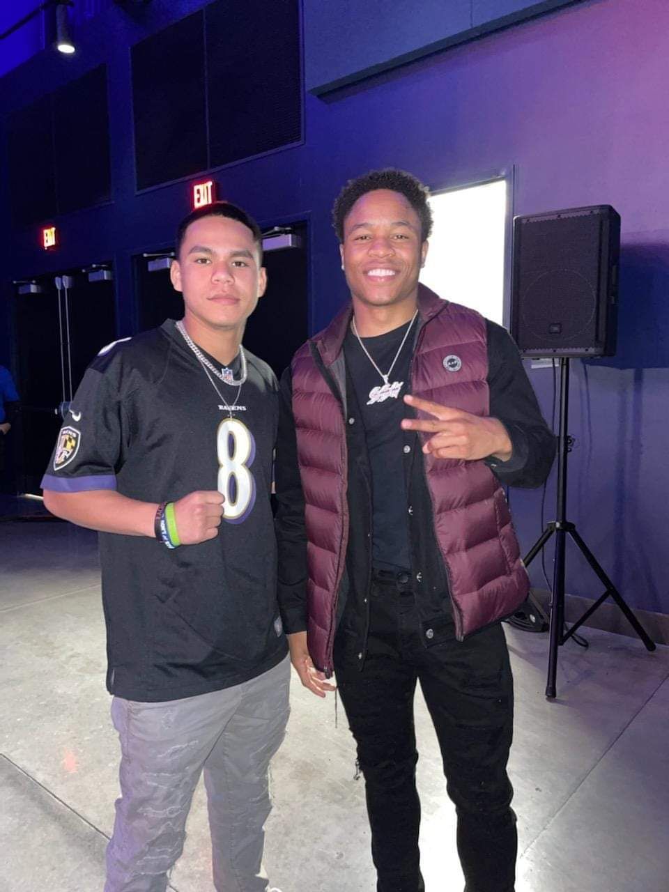 Two people posing indoors, one in a black jersey and one in a burgundy vest, with a purple-lit wall behind them