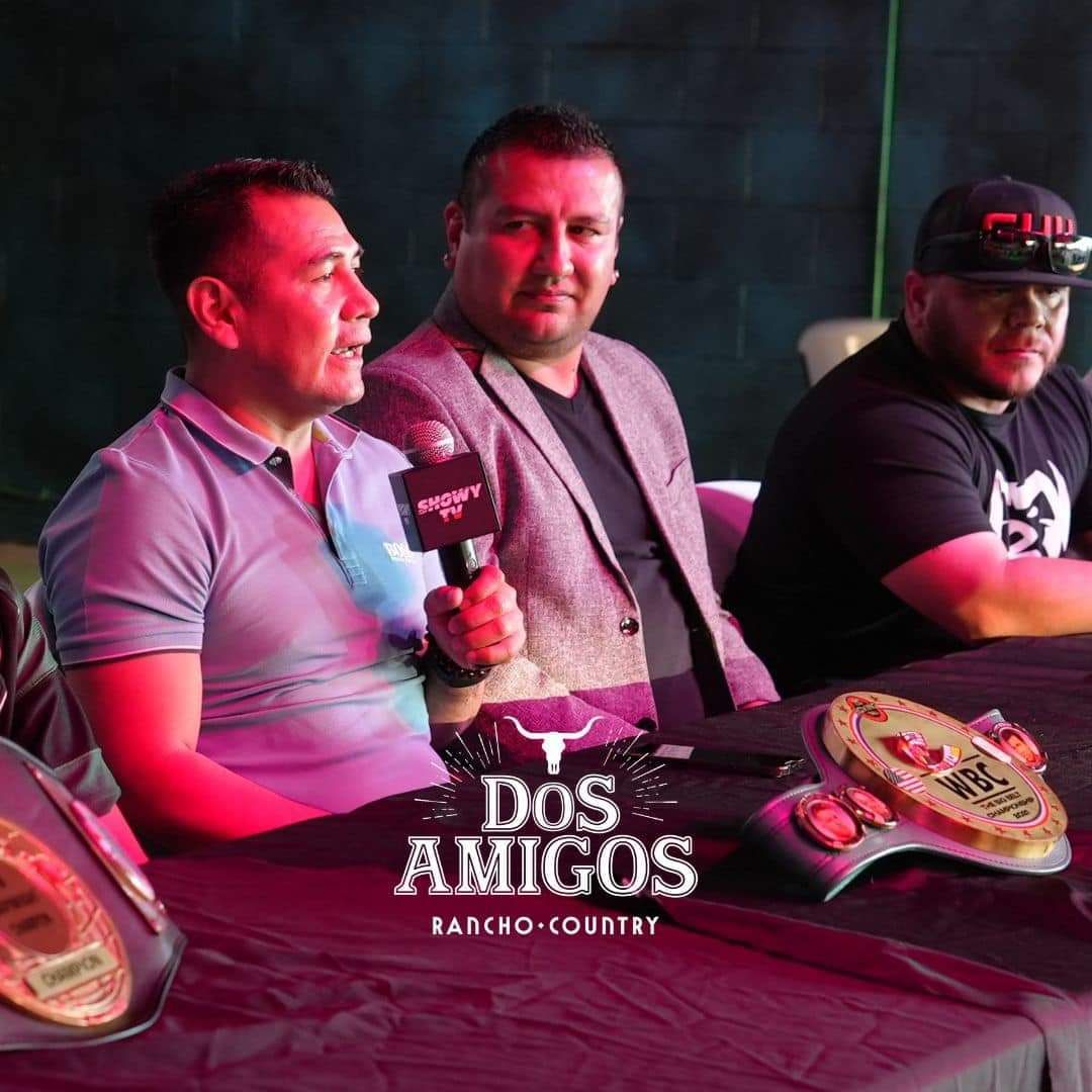 Three men at a table with championship belts, lit by pink stage lighting, in a Dos Amigos Ranch Country setting