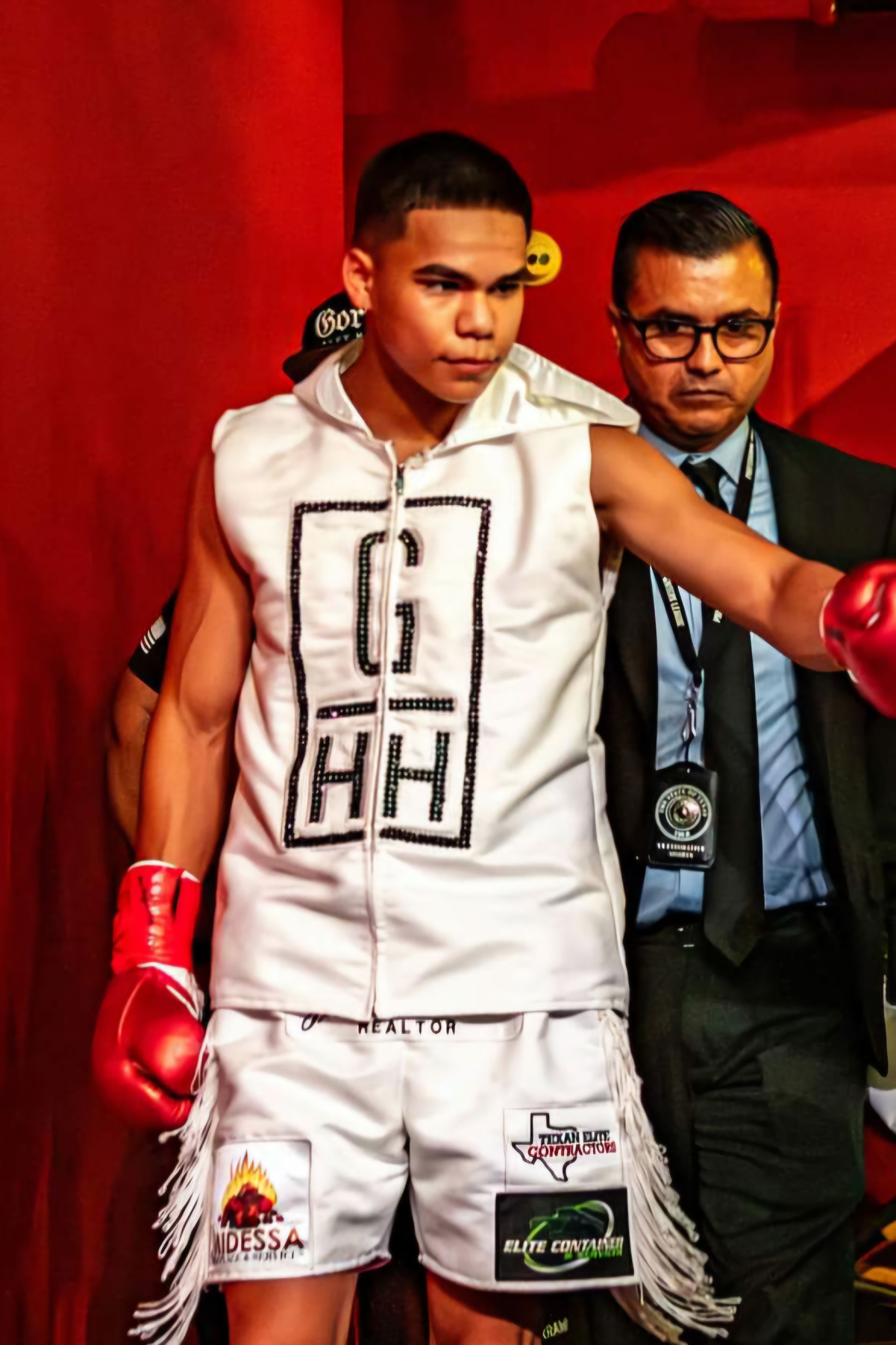 Boxer in white robe and red gloves stands in a red-lit ring with a man in a suit behind him.