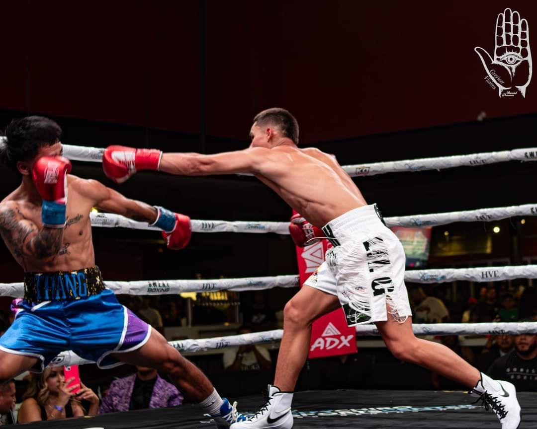 Boxers spar in a ring, one in white shorts jabbing a crouched opponent in blue shorts.