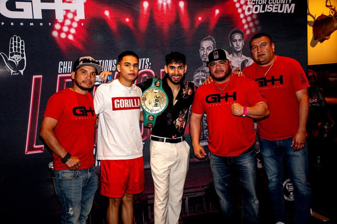 Five men pose in a sports venue, one holding a championship belt, with red-and-black event banners behind them.