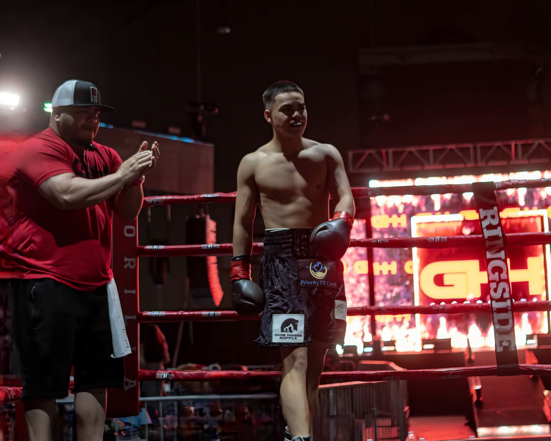 Two men in a boxing ring, one shirtless in gloves, with bright red lights and a coach applauding nearby