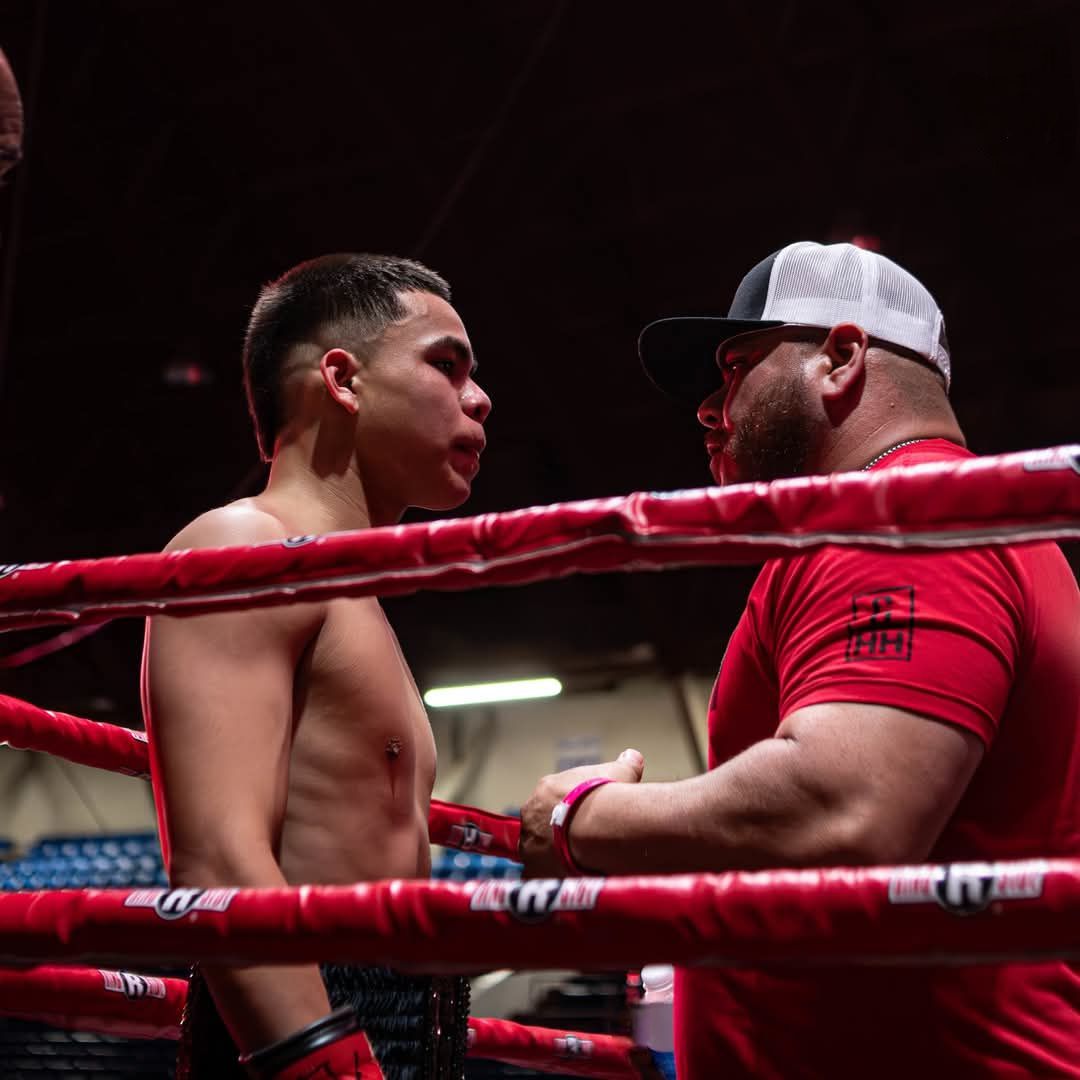 Two boxers face off in a ring, with red ropes and one wearing a red shirt and white cap.