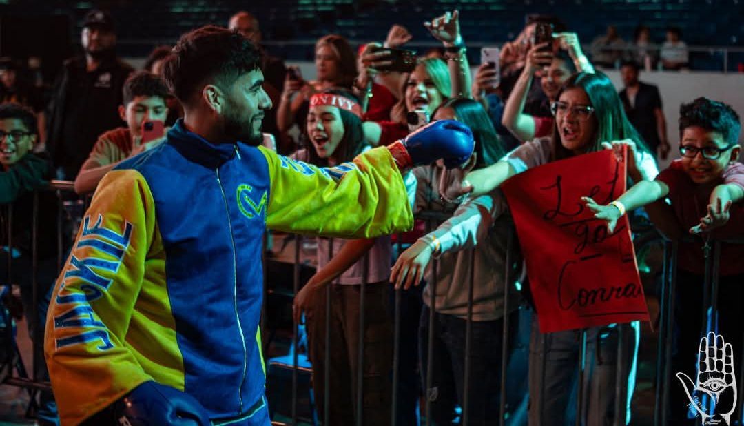 Man in blue and neon jacket gesturing to cheering crowd behind barricade at night.