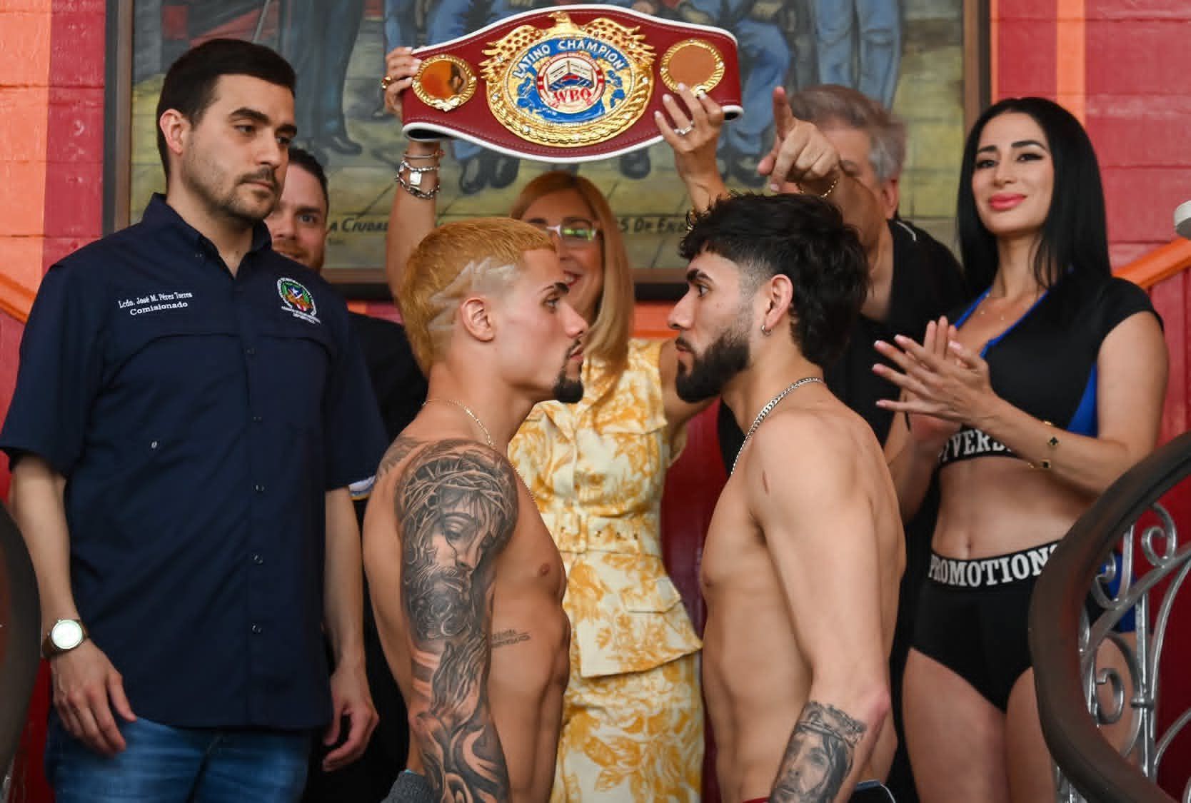 Two boxers face off at a weigh-in with a championship belt behind them, surrounded by officials and spectators.