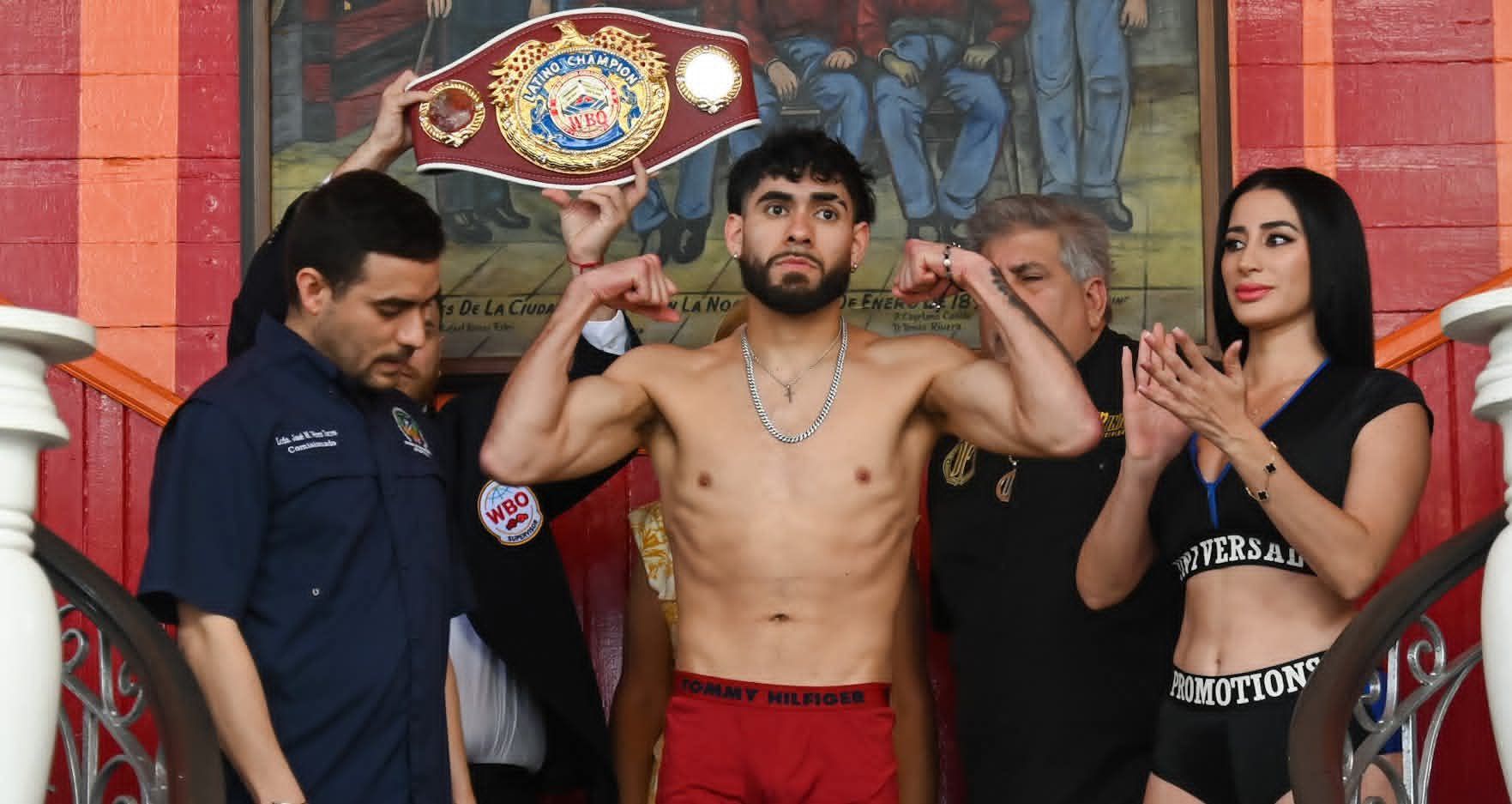 Shirtless boxer poses with arms raised in a ring, flanked by two people, with a championship belt behind him.