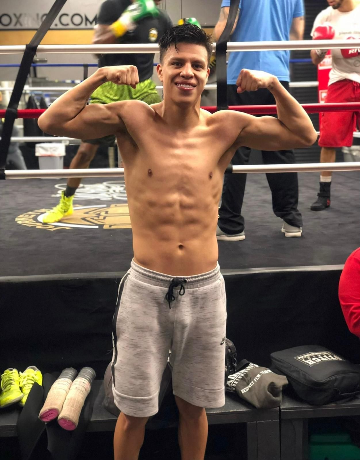 Boxer posing in a gym ring, flexing both arms and smiling, with training gear visible nearby.