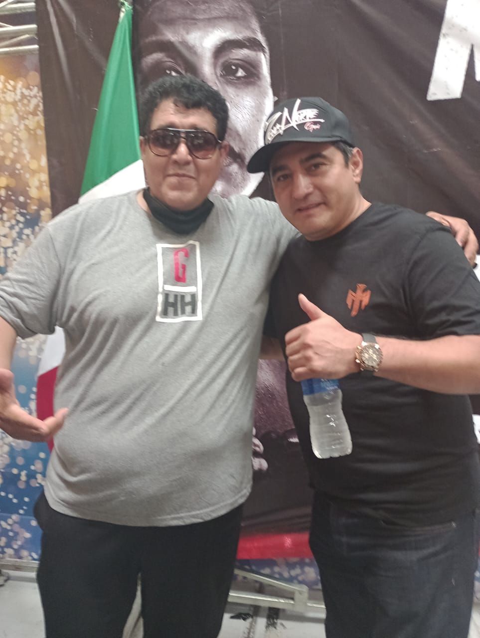 Two men pose together indoors, one giving a thumbs-up, with a large face poster behind them.