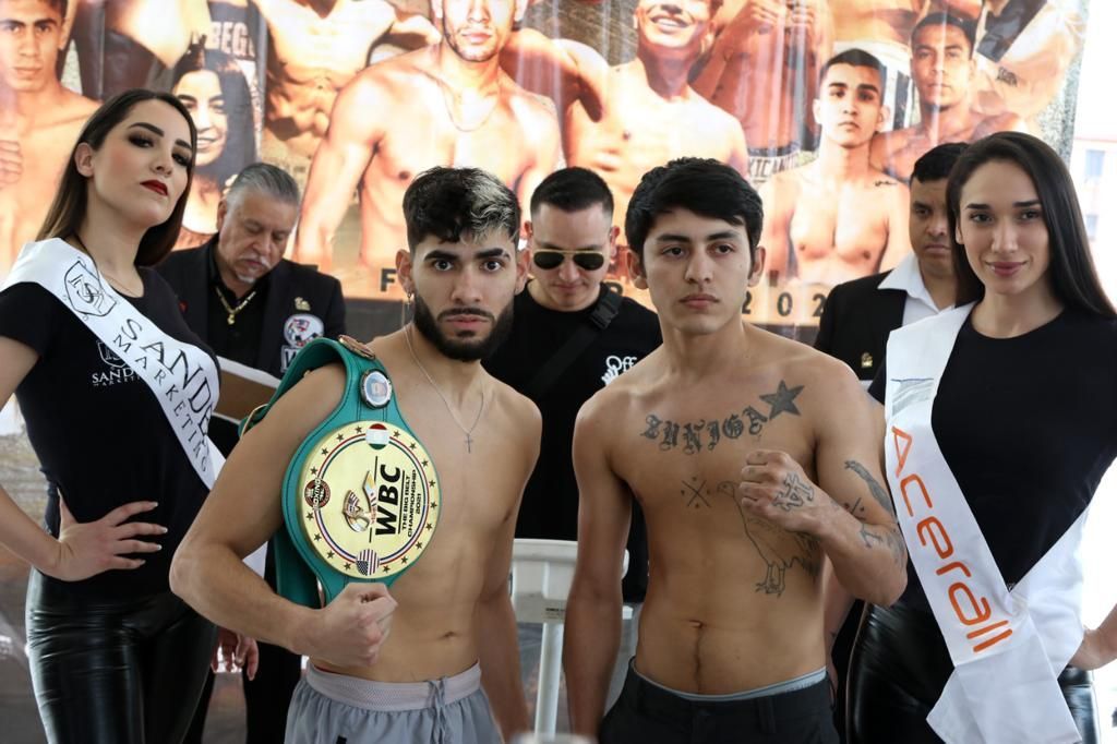 Two shirtless fighters with a championship belt stand beside two sash-wearing women at a boxing event.