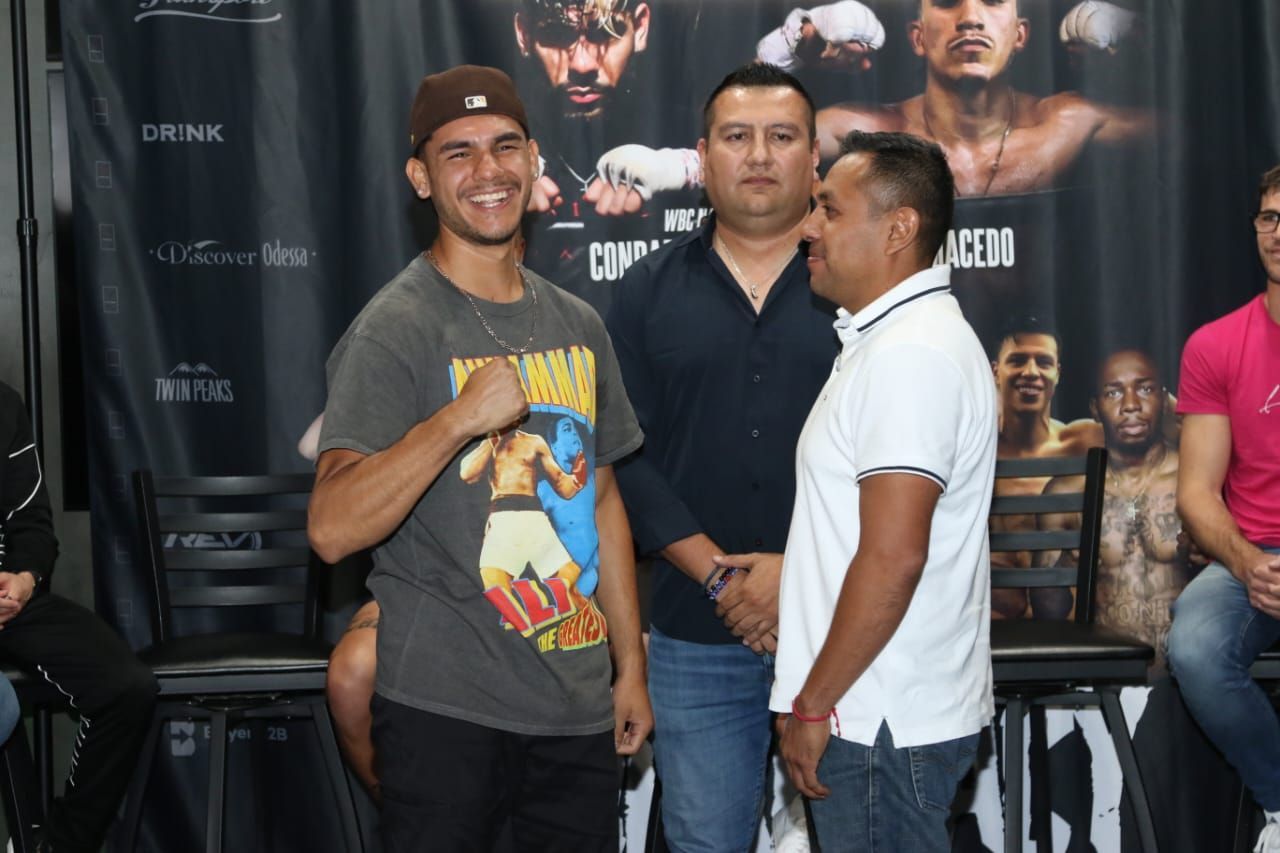 Three men posing at a boxing event backdrop, one smiling and giving a thumbs-up.