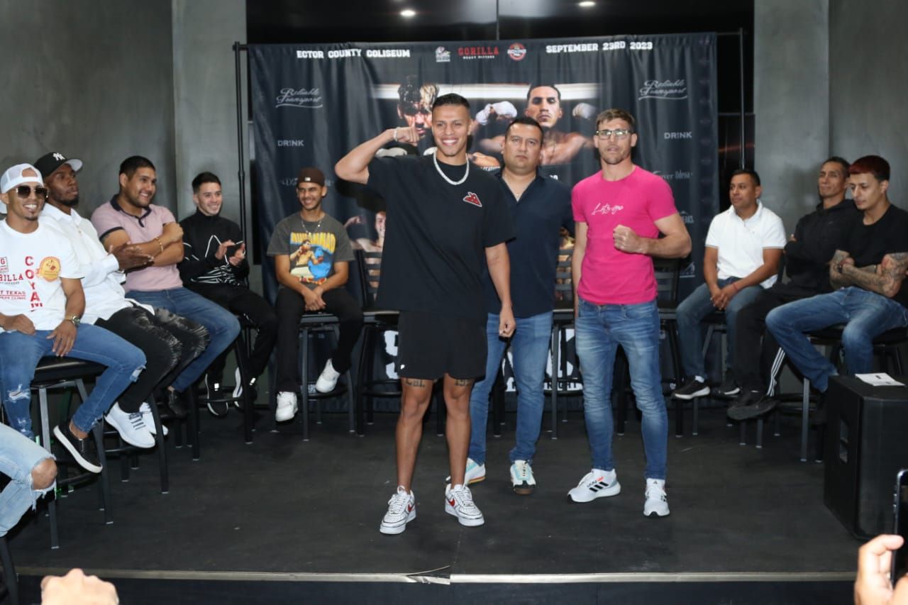 Two men stand on a stage before an audience, one saluting in a pink shirt and one in black