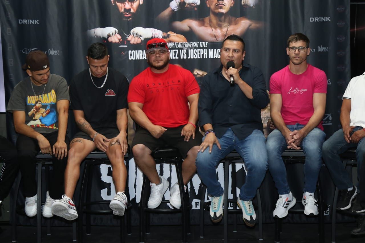 Five men seated on a stage panel, one speaking into a microphone, with event banners behind them.