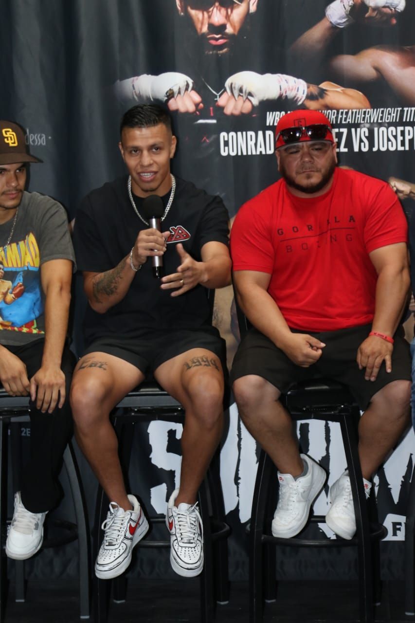 Three men seated on stools at a panel event, one speaking into a microphone, with a large backdrop behind them.