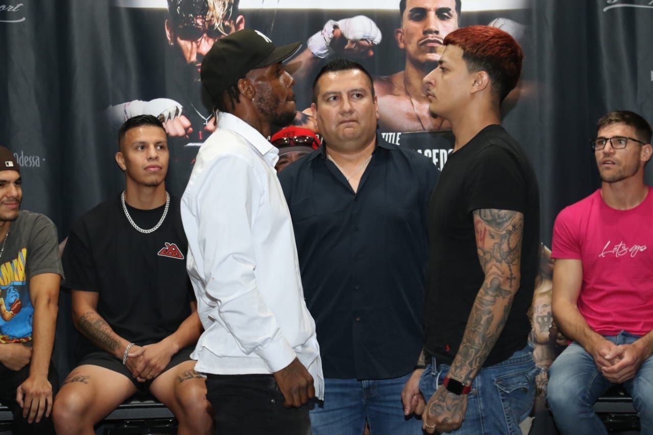 Two men face off at a press event, with seated spectators and a large poster backdrop behind them.