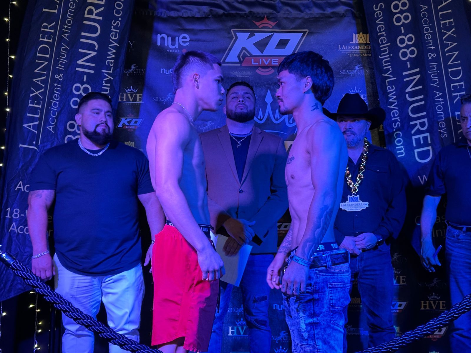 Five men posing in a blue-lit nightclub, with one shirtless man in red shorts facing another man.