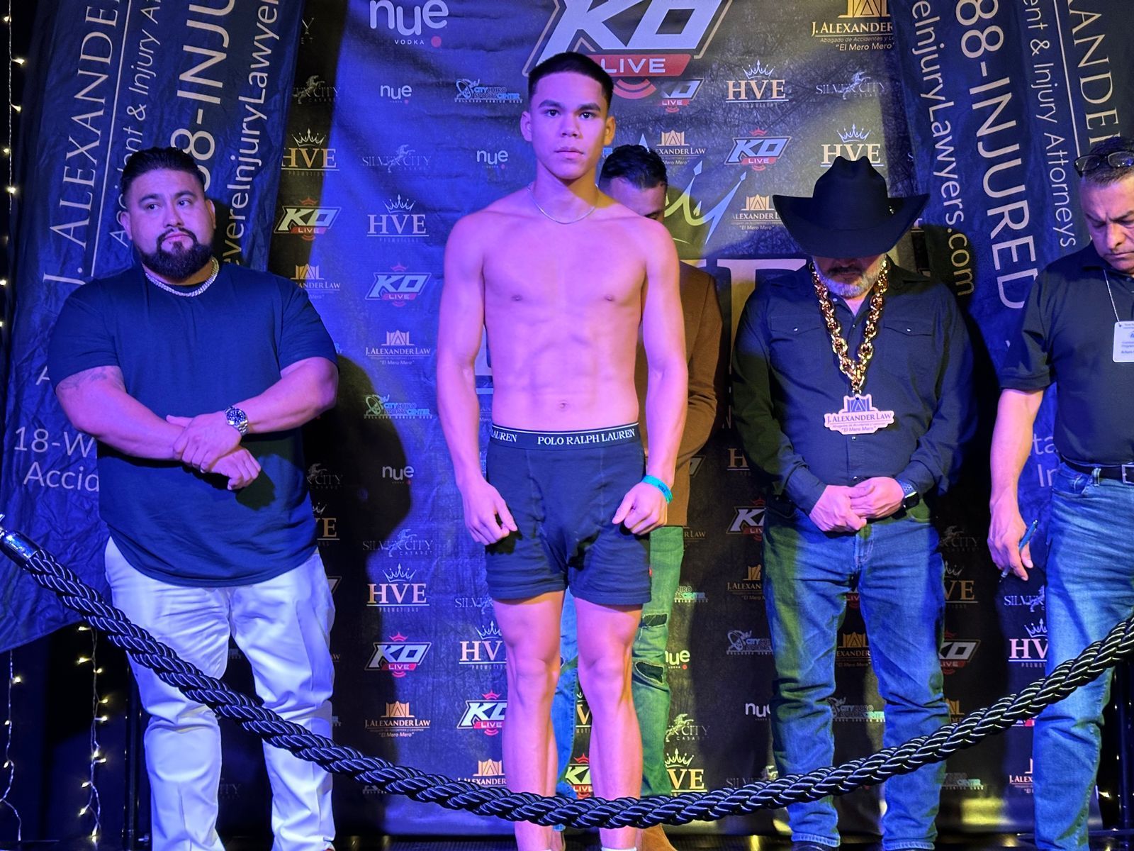 Three men posing on a purple event stage with a boxing-style backdrop and rope barrier