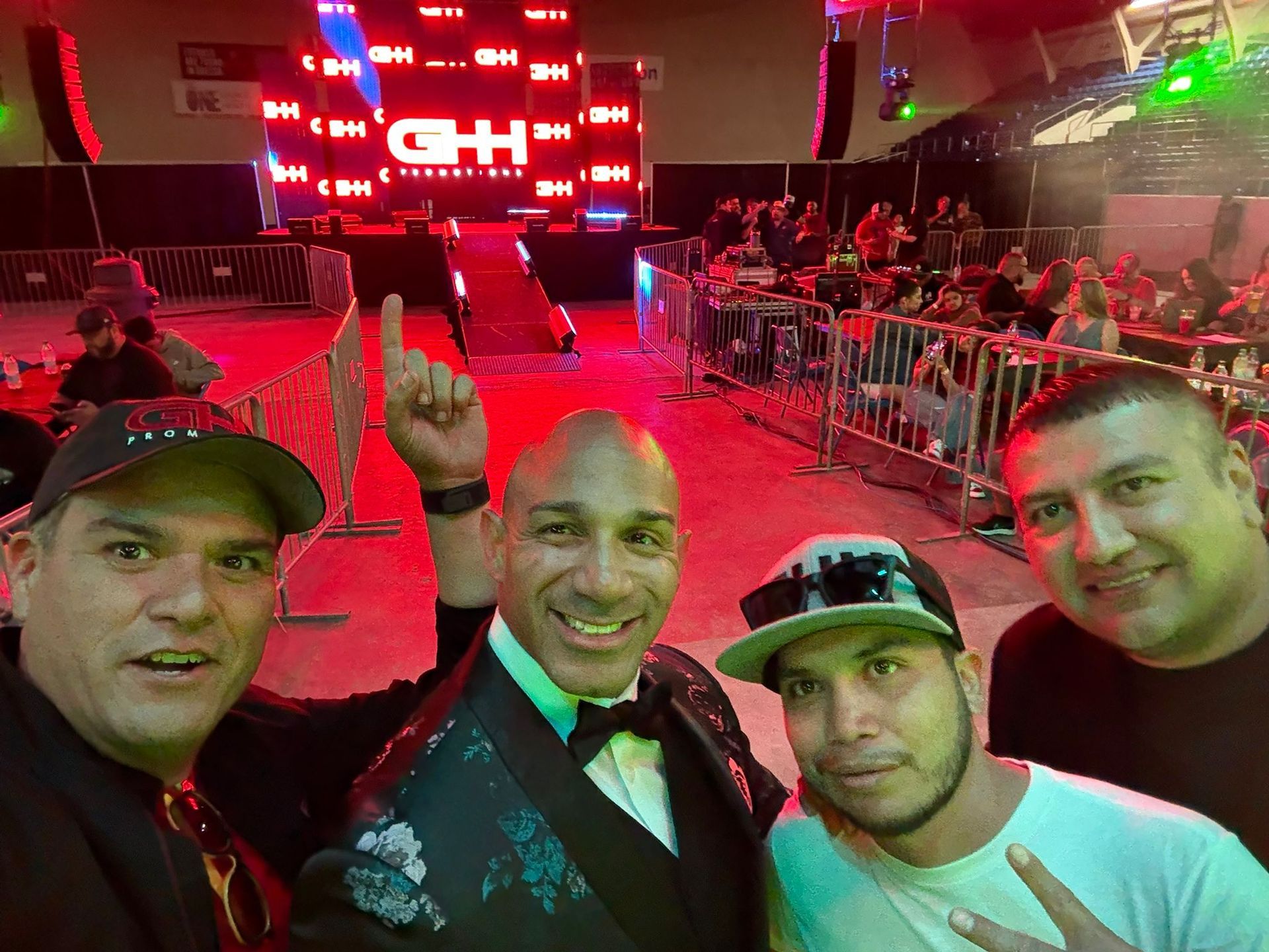 Four men posing for a selfie in a red-lit concert arena with a stage in the background