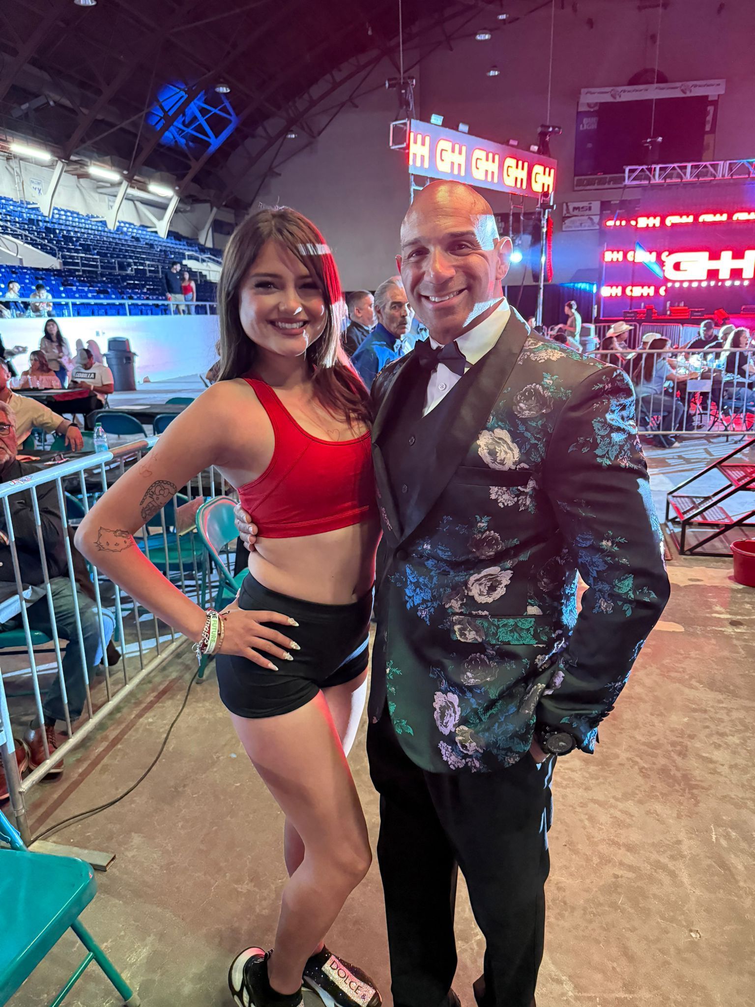 Two people posing at a lively event, the woman in a red top and black shorts, the man in a patterned jacket.