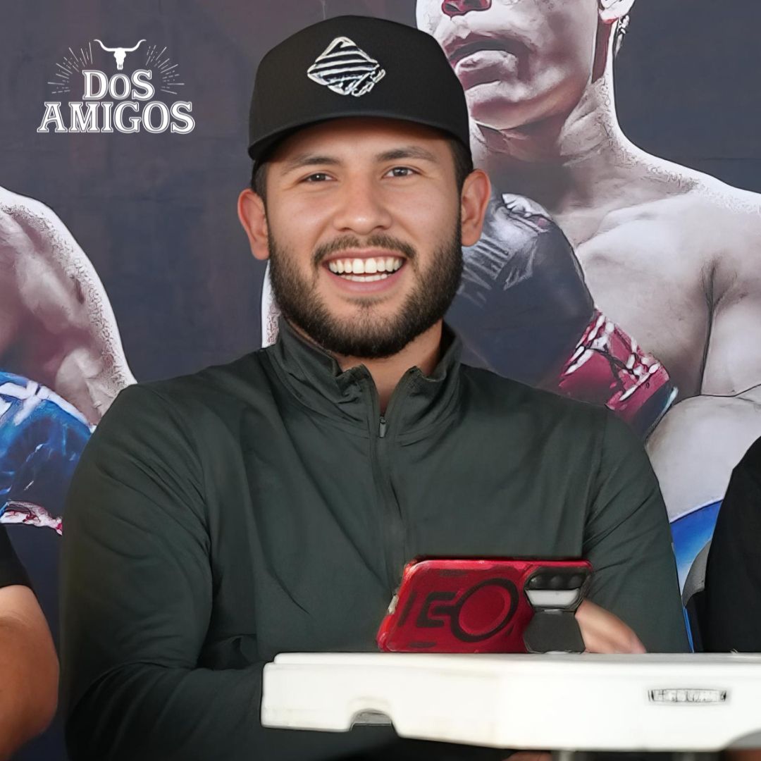 Smiling man in black cap and jacket holding a red item at a Dos Amigos press event backdrop
