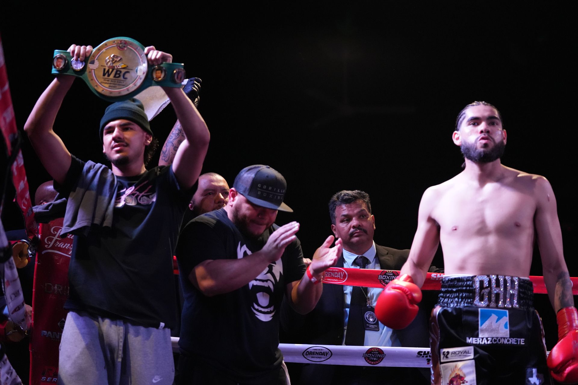 Boxer in red gloves stands in ring beside team member holding championship belt overhead
