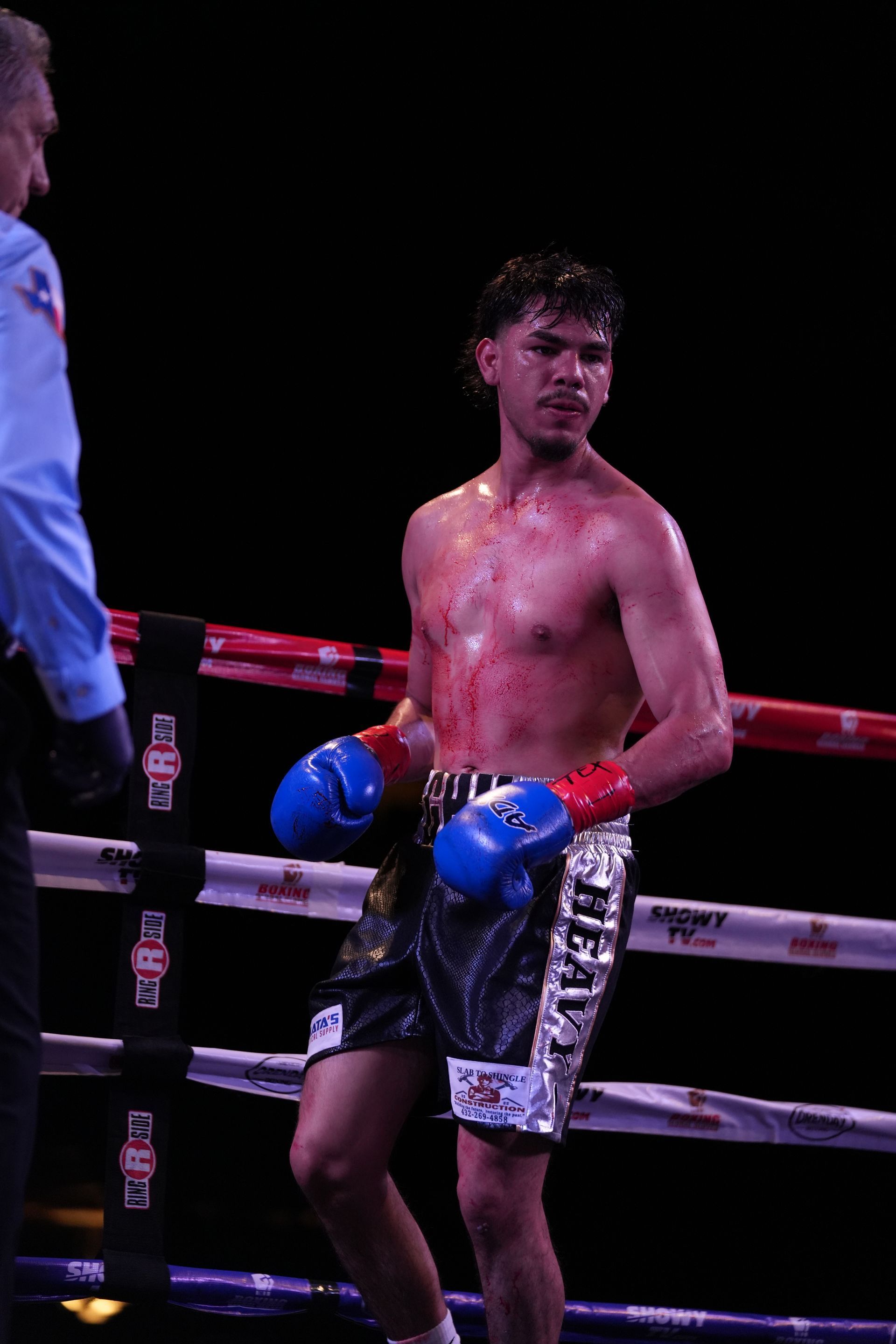 Boxer in blue gloves leaning on the ropes in a ring, shirtless and glistening with sweat under purple lights