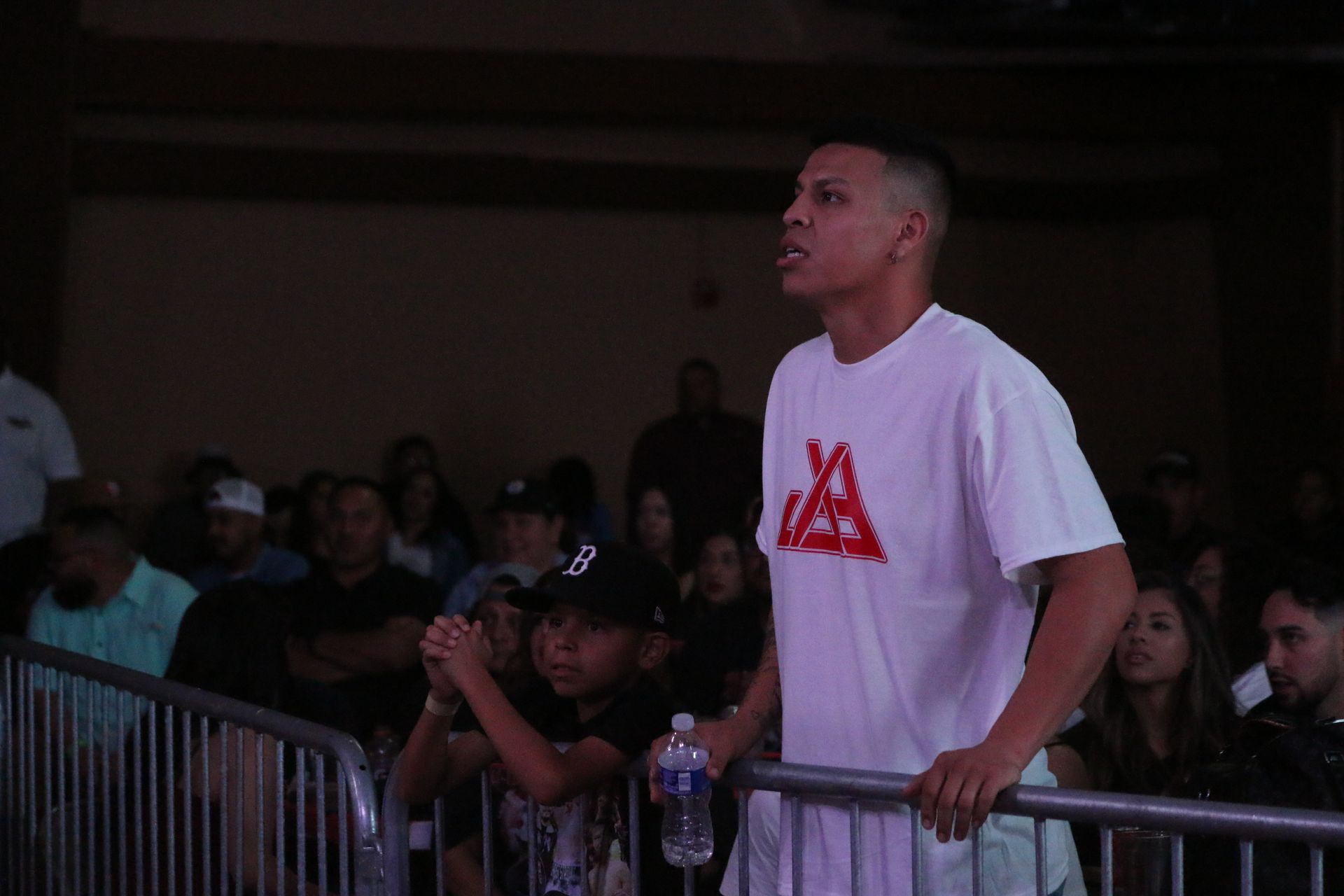 Man in a white T-shirt leans on a barrier, watching an event in a dim crowd.