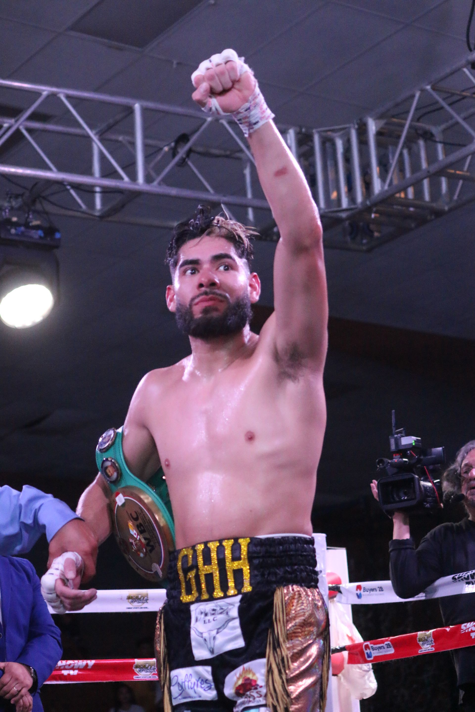 Boxer in a ring raising a gloved fist in victory, with a camera crew behind him.