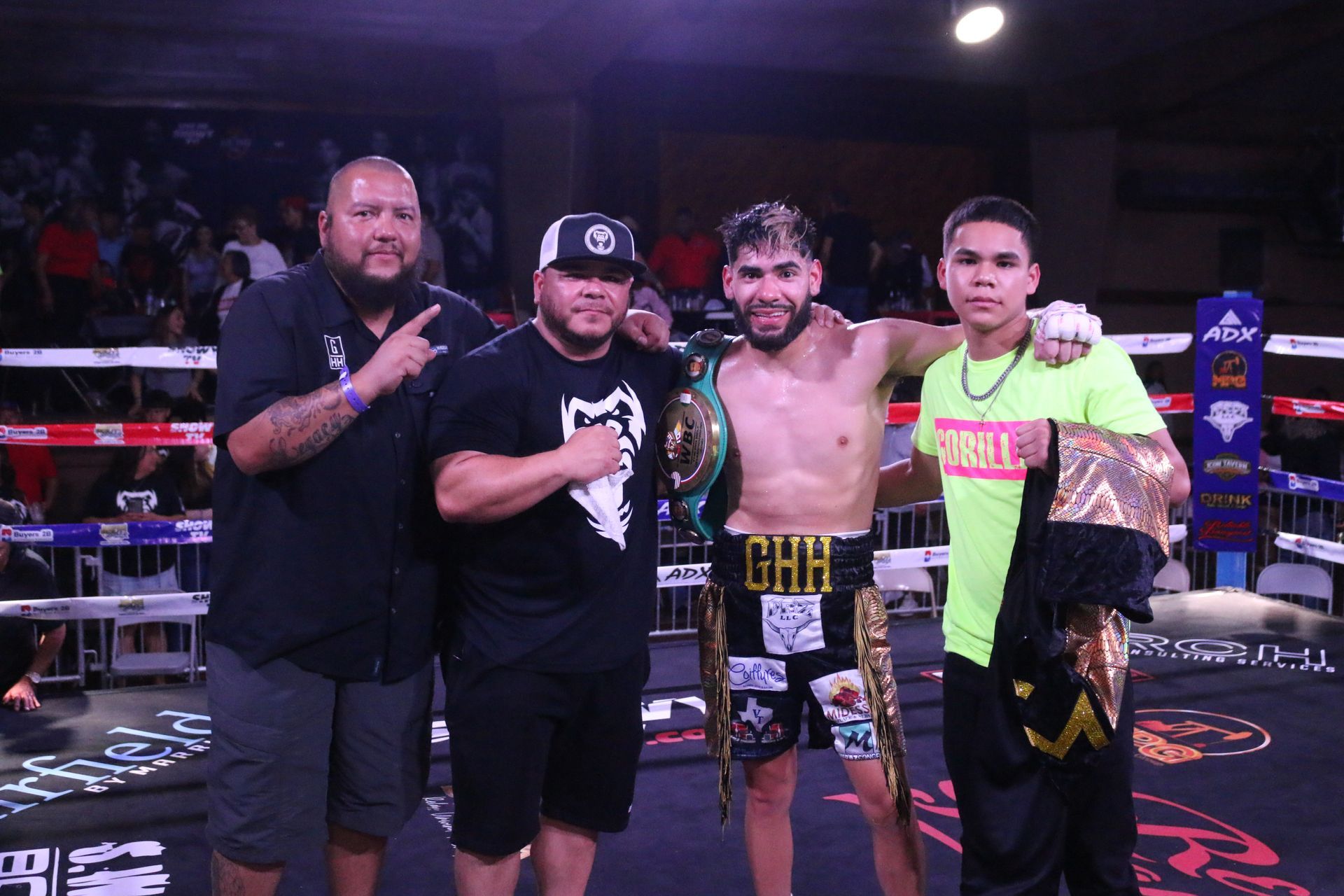 Four people pose in a boxing ring, including a shirtless champion holding a belt and trophy.