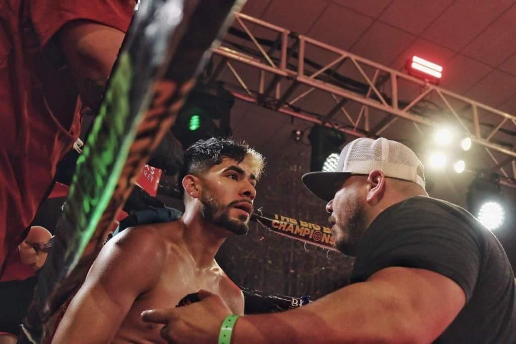 Two men in a cage fight, one in black and white headgear holding the other at close range under bright arena lights