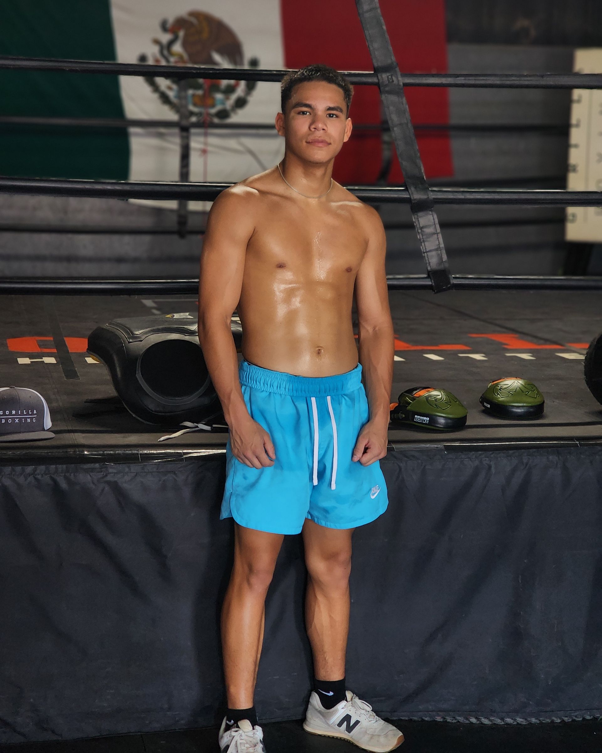 Boxer standing in a ring wearing blue shorts and white sneakers, with gloves on the ropes behind him.