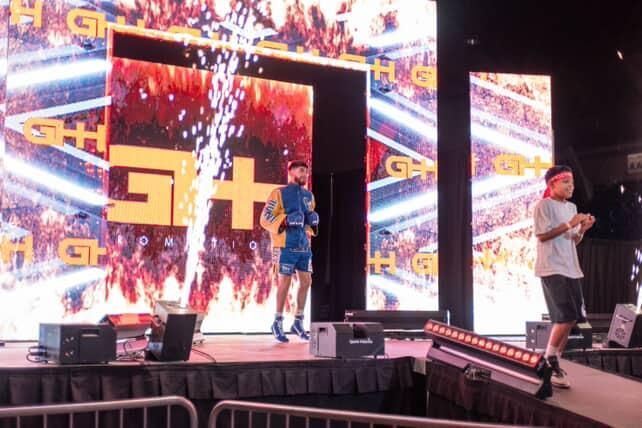 Two people on a stage in front of bright LED screens with gold graphics and fireworks-like effects