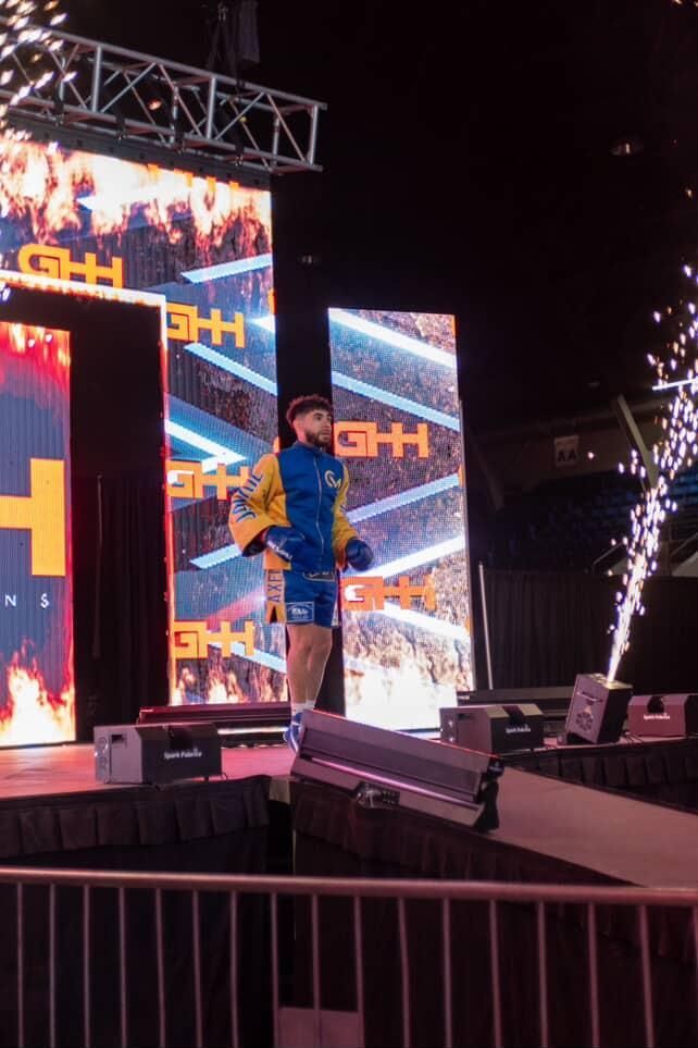 Person in blue outfit standing on a brightly lit stage with LED screens and fireworks.