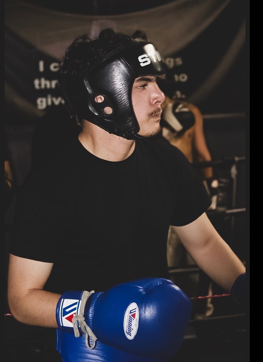 A person wearing black boxing gear and blue gloves stands in a boxing gym with a serious expression.