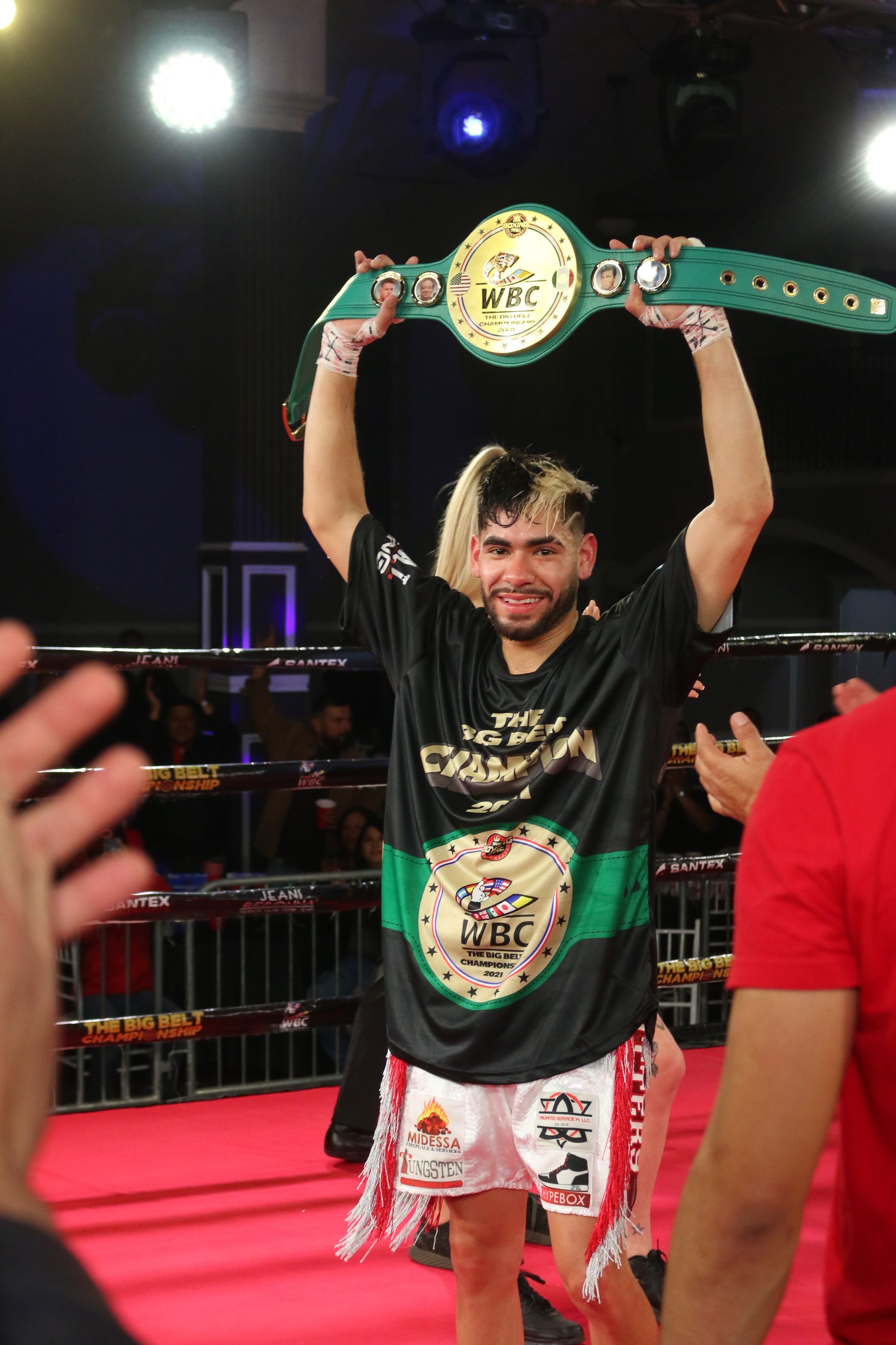 Boxer holding a championship belt overhead in a ring, wearing black and green gear, with crowd lights behind him