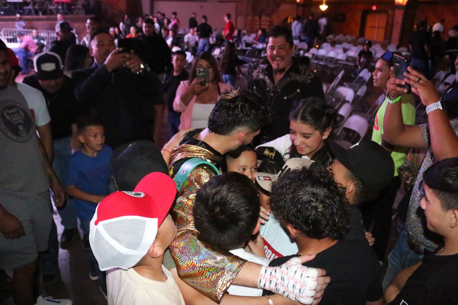 People crowd around a table at a lively indoor event, leaning in and talking under pink lighting.